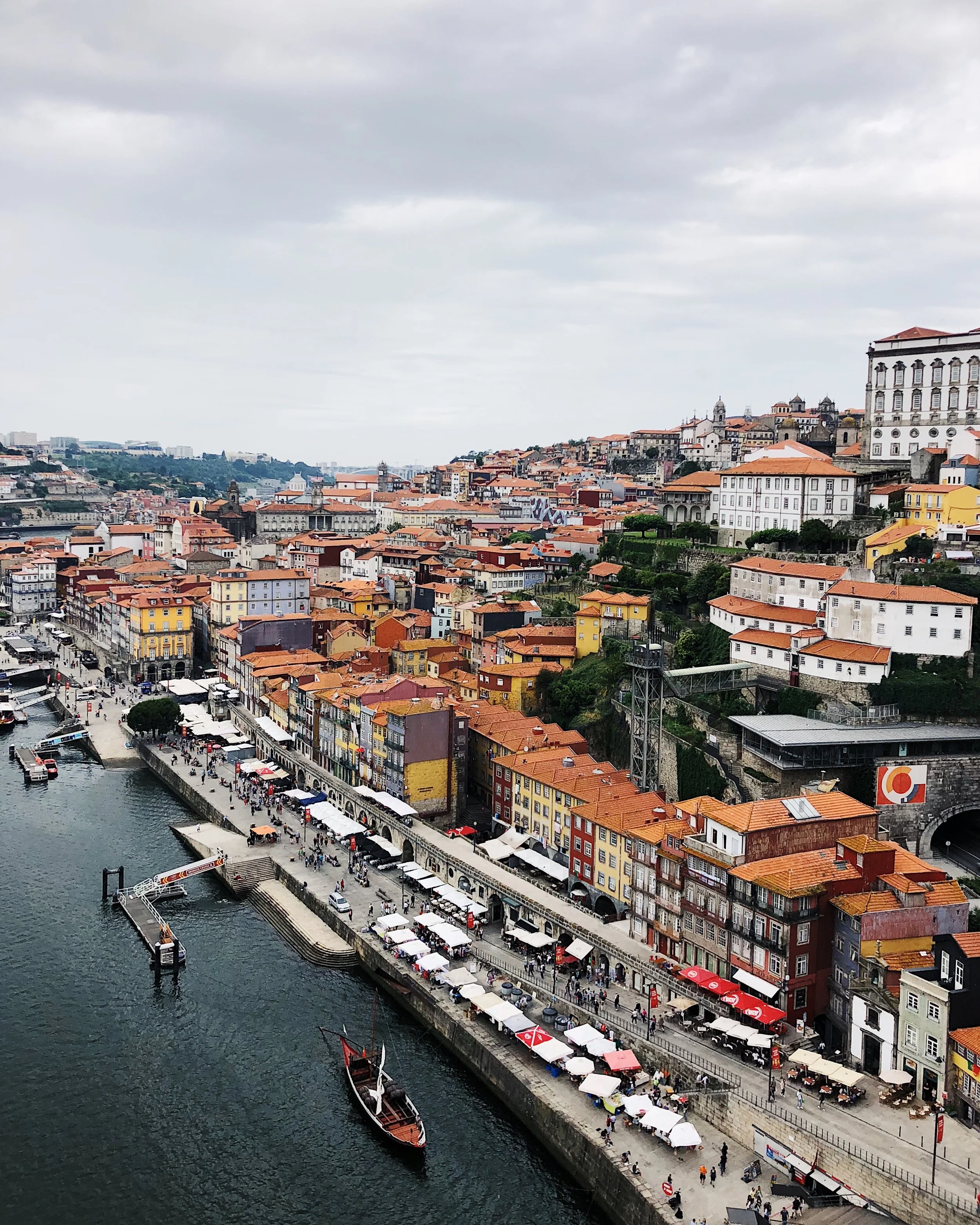 A scenic view of a European city with colorful buildings along a river, outdoor cafes with white umbrellas, and a boat docked in the water. Hills with more buildings and a cloudy sky are in the background.
