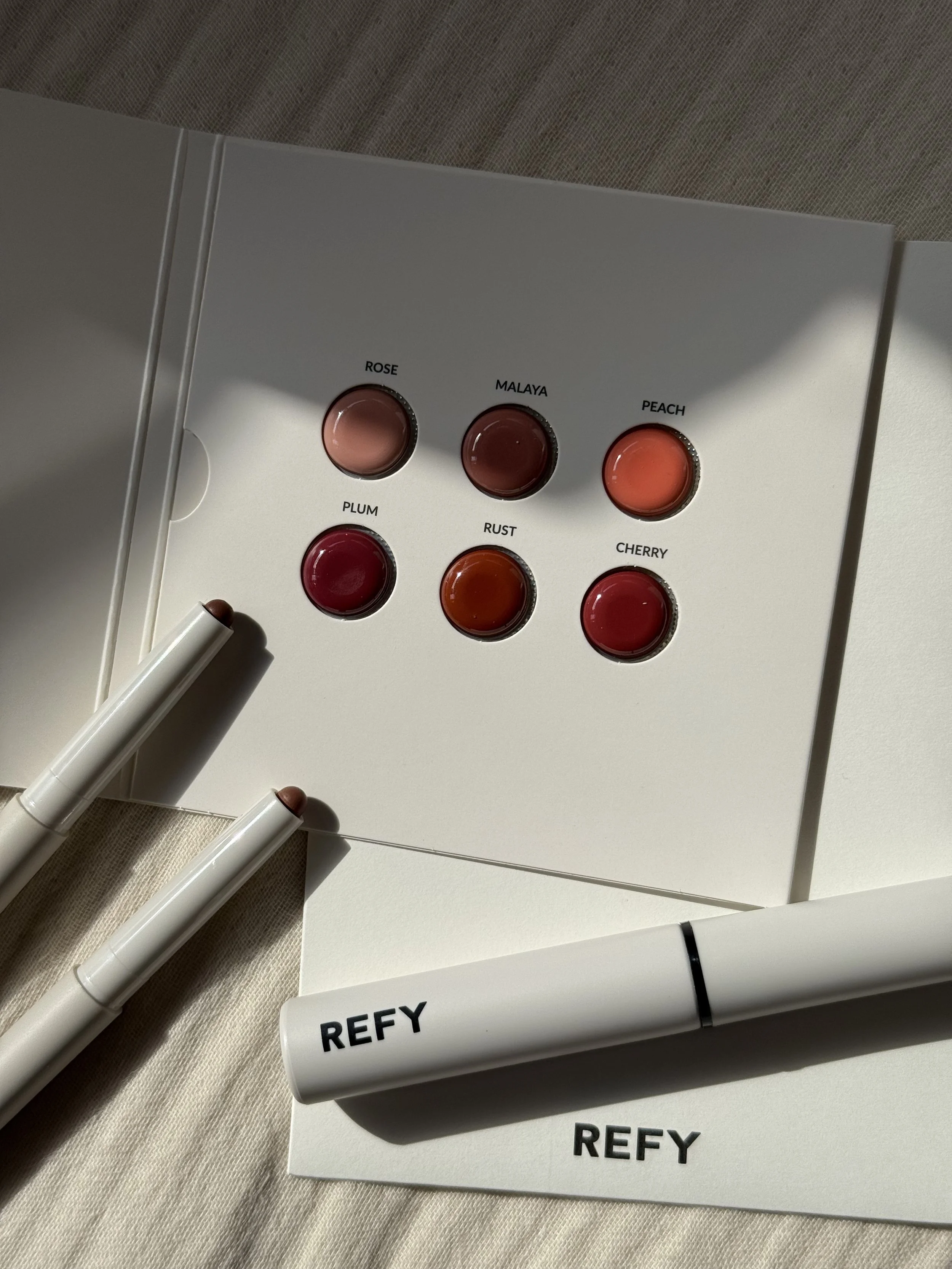 Color swatch and lip products from Refy on a white surface, with a color palette showing shades named Rose, Malaya, Peach, Plum, Rust, and Cherry.