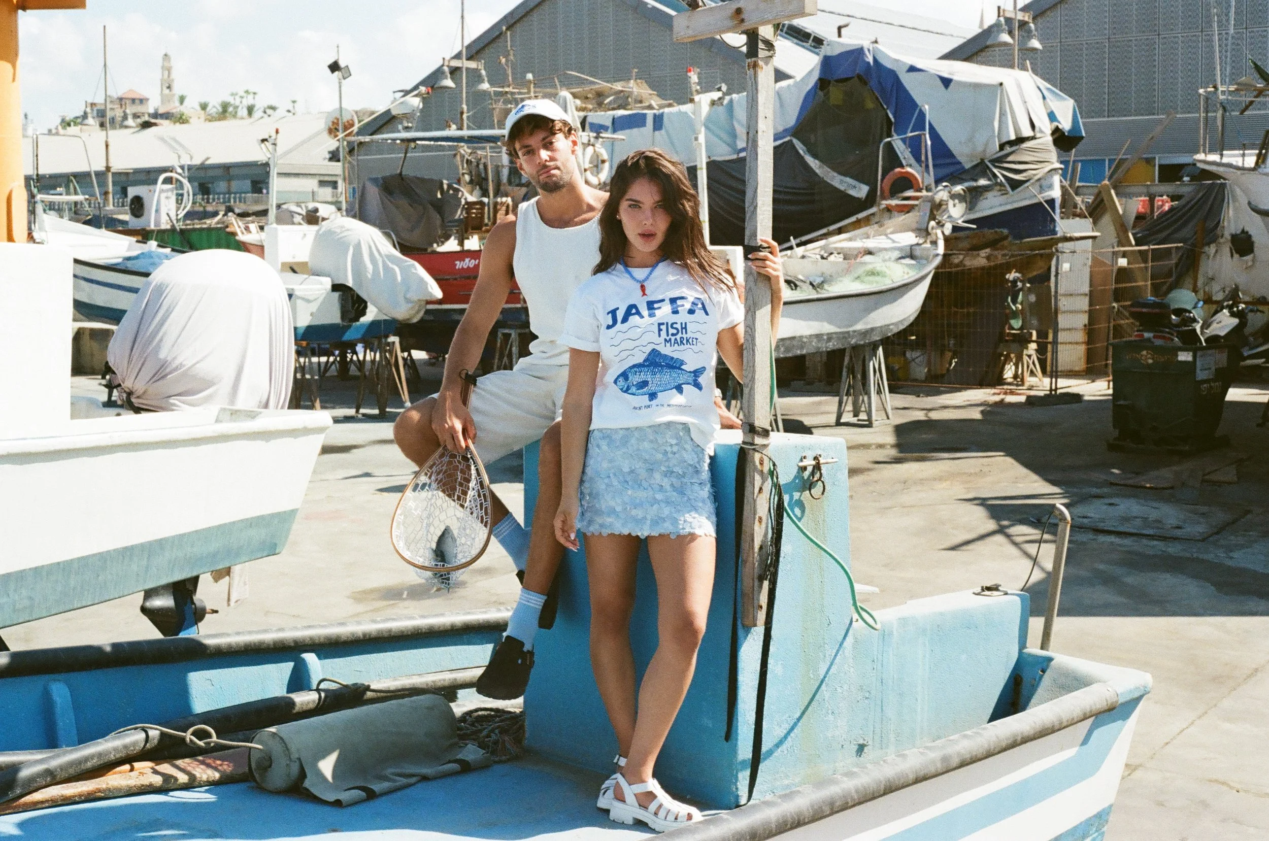 Two young people standing on a small boat at a marina with boats and fishing equipment in the background.