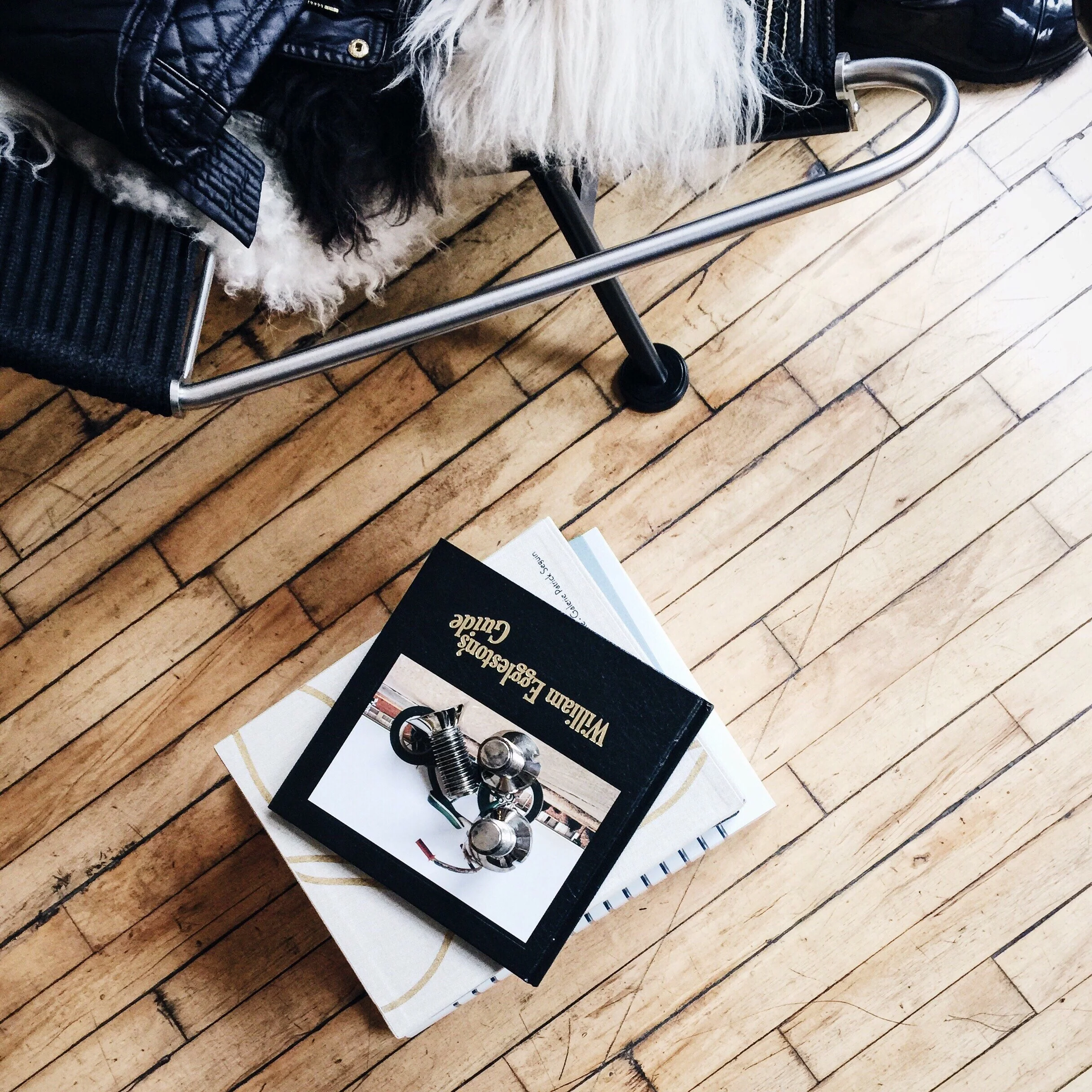 A view of a wooden floor with a stack of books, one titled 'William Egeston’s Guide', and a pair of metallic shaped objects on top of the books. Part of a black leather jacket and a dog with curly fur lying on a chair are also visible.