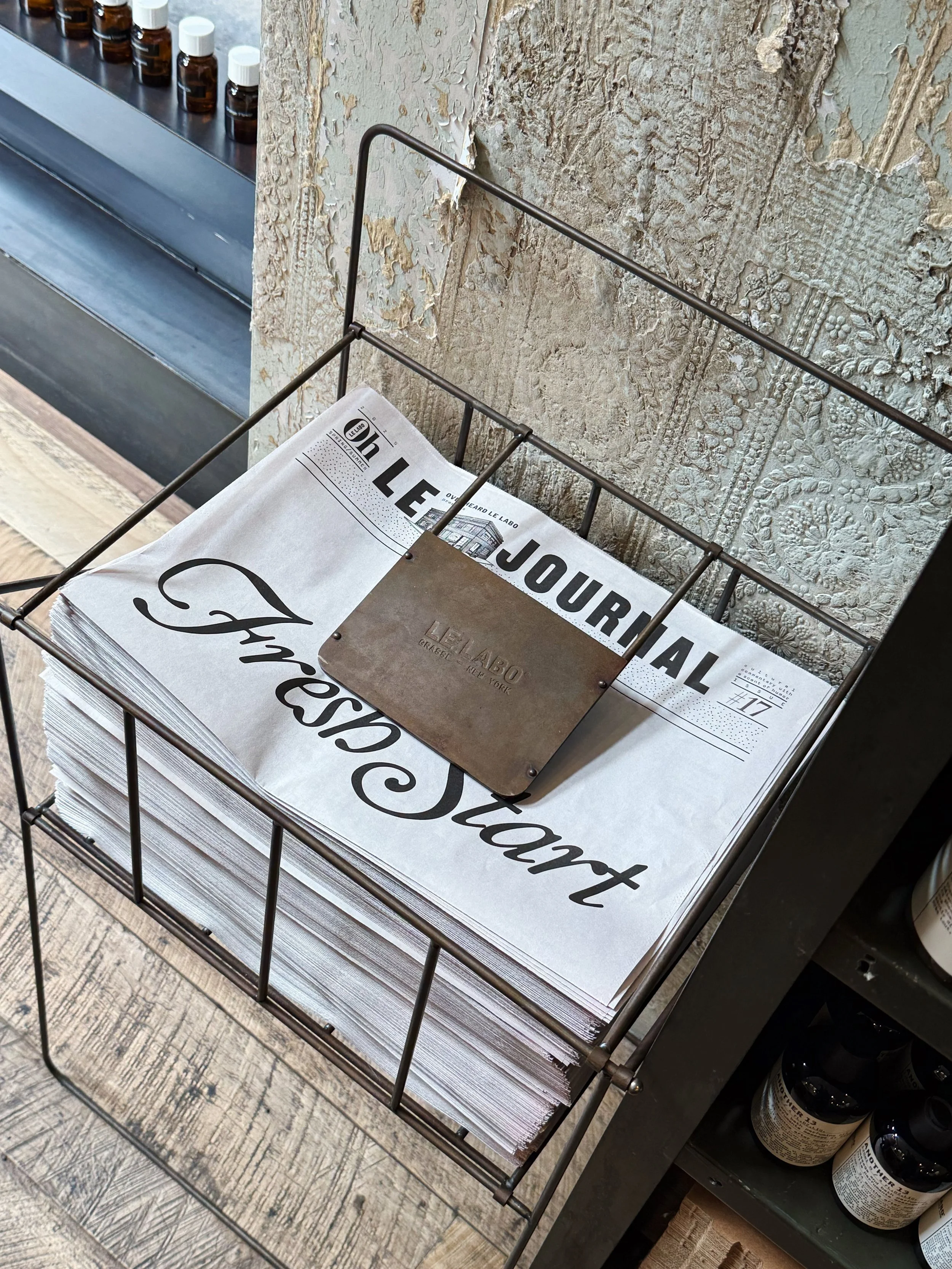 A wire magazine rack holding newspapers, with a decorative metal plate on top, positioned against a textured wall. In the background, part of a shelf with small bottles is visible.