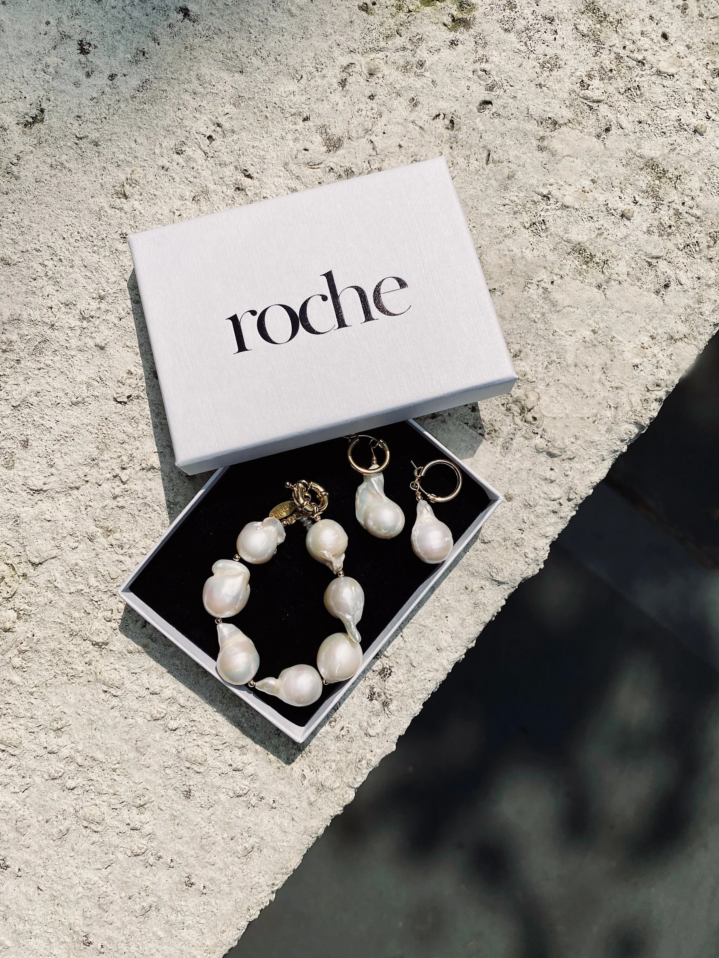 A white box labeled 'roche' is open, revealing pearl jewelry including a bracelet and earrings, placed on a textured light-colored surface.