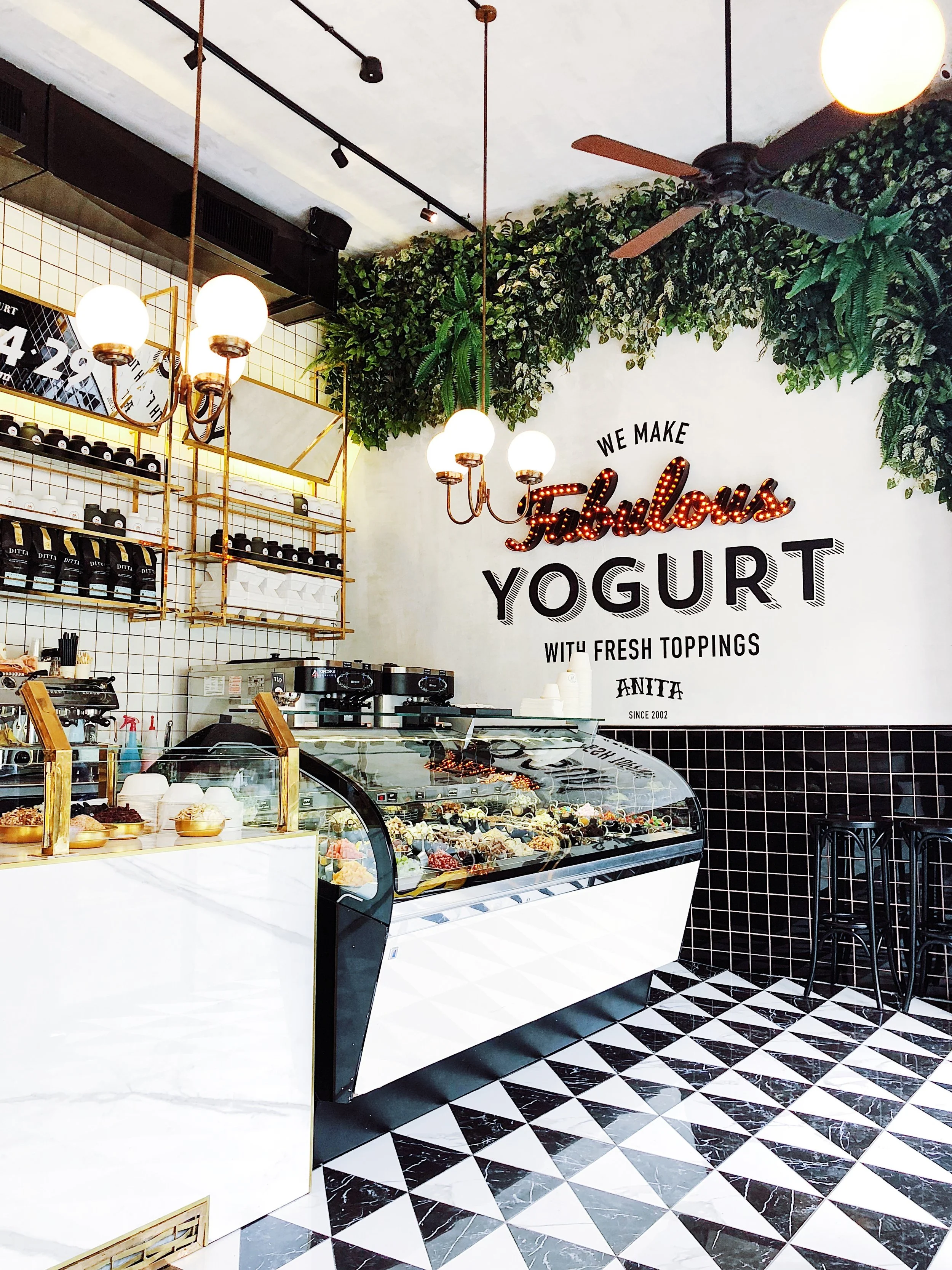Inside a modern yogurt shop with a black and white tiled floor, a decorated ice cream display case, and a wall with the words 'We make fabulous yogurt with fresh toppings'. The shop has hanging globe lights, green wall plants, and black bar stools.