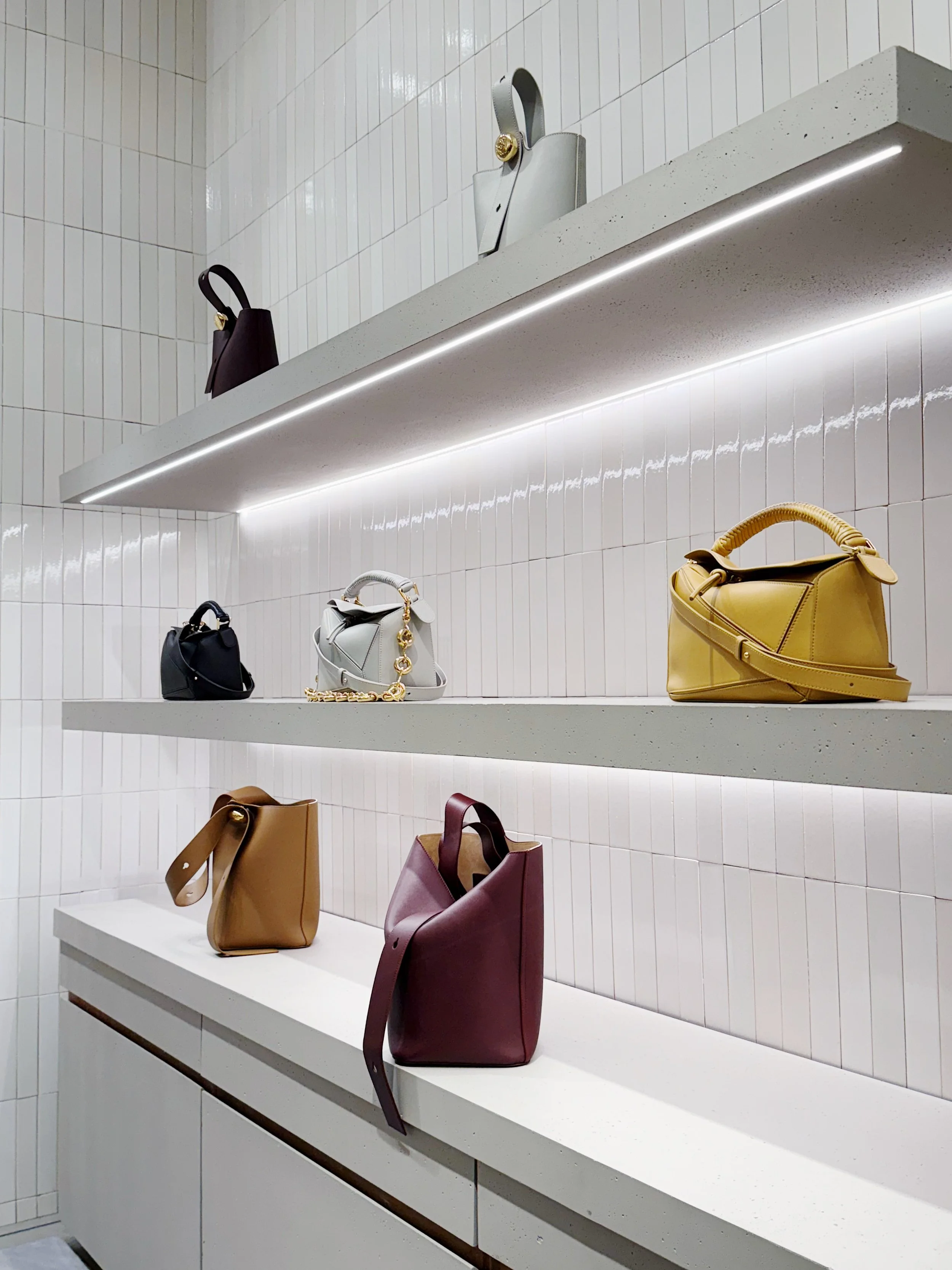 Display of small handbags on minimalist shelves in a retail store with white tiled walls and subtle lighting.