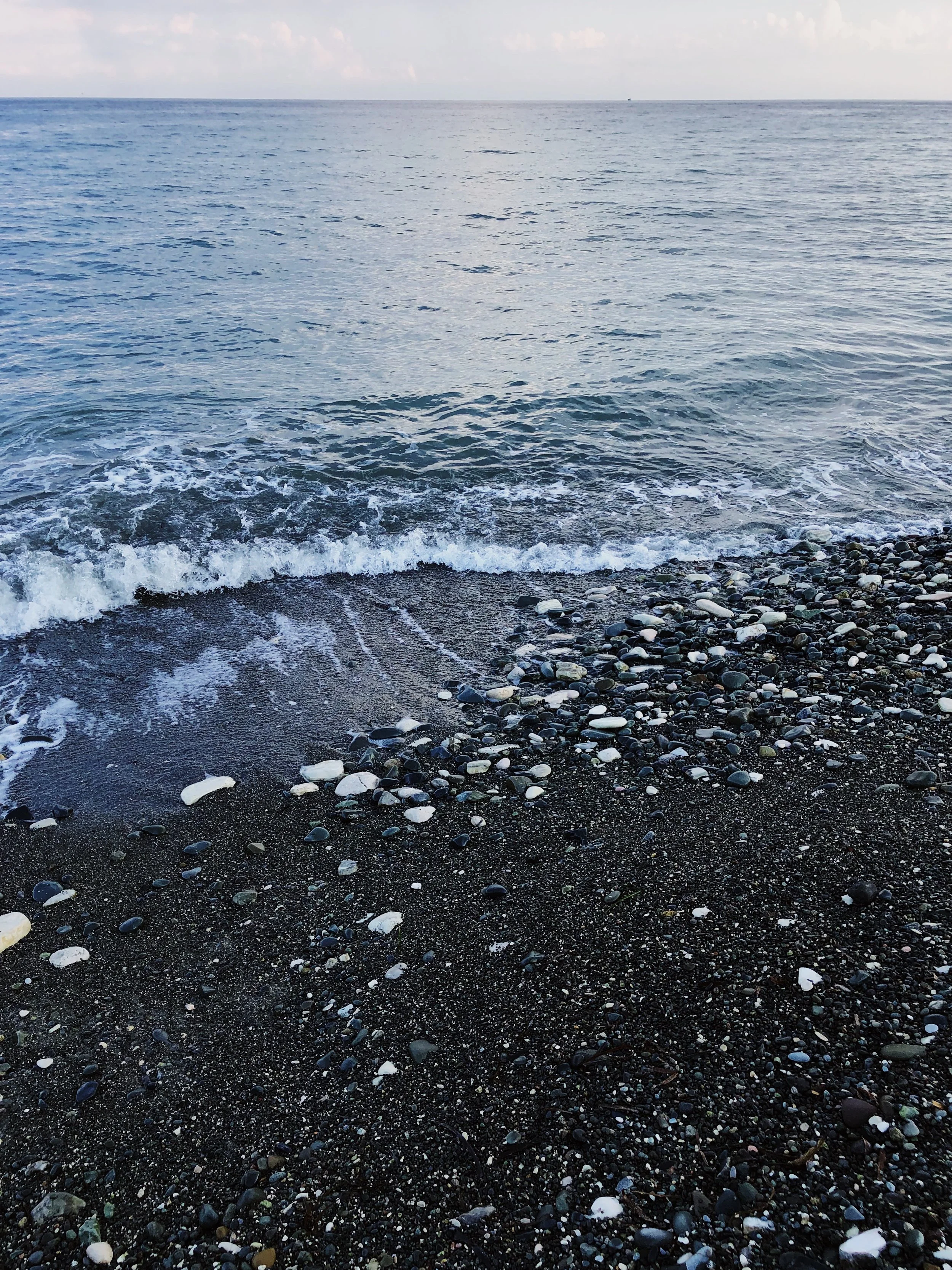 Black pebble beach with small rocks and ocean waves, calm sea, cloudy sky.
