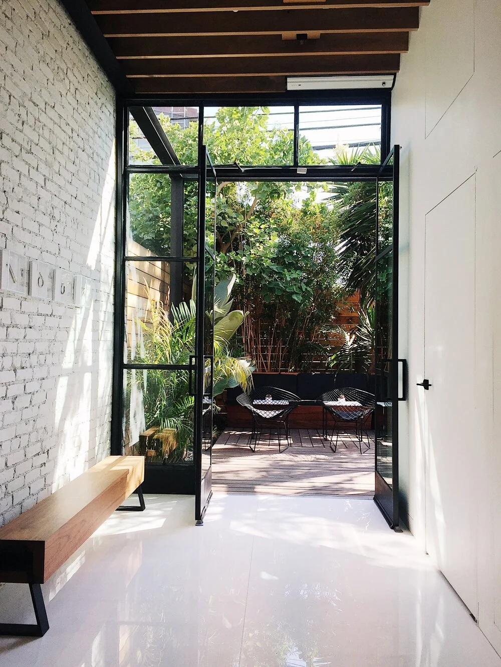 A modern living space with white brick wall, wooden beam ceiling, and open glass door leading to a small outdoor patio with lush green plants and two black wire chairs.