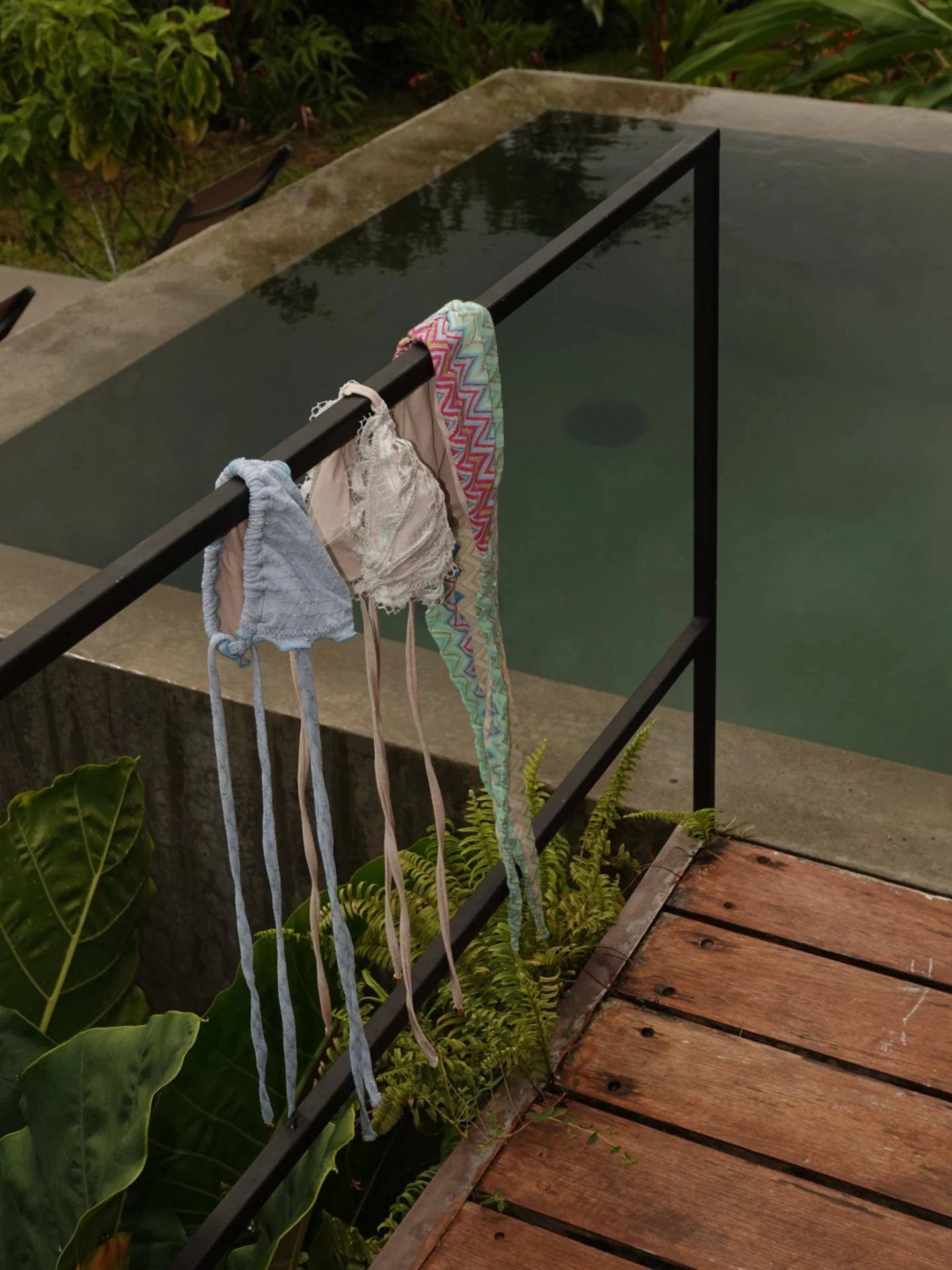 Colorful swimsuits hanging on a black railing next to a swimming pool, with green plants and wooden decking nearby.