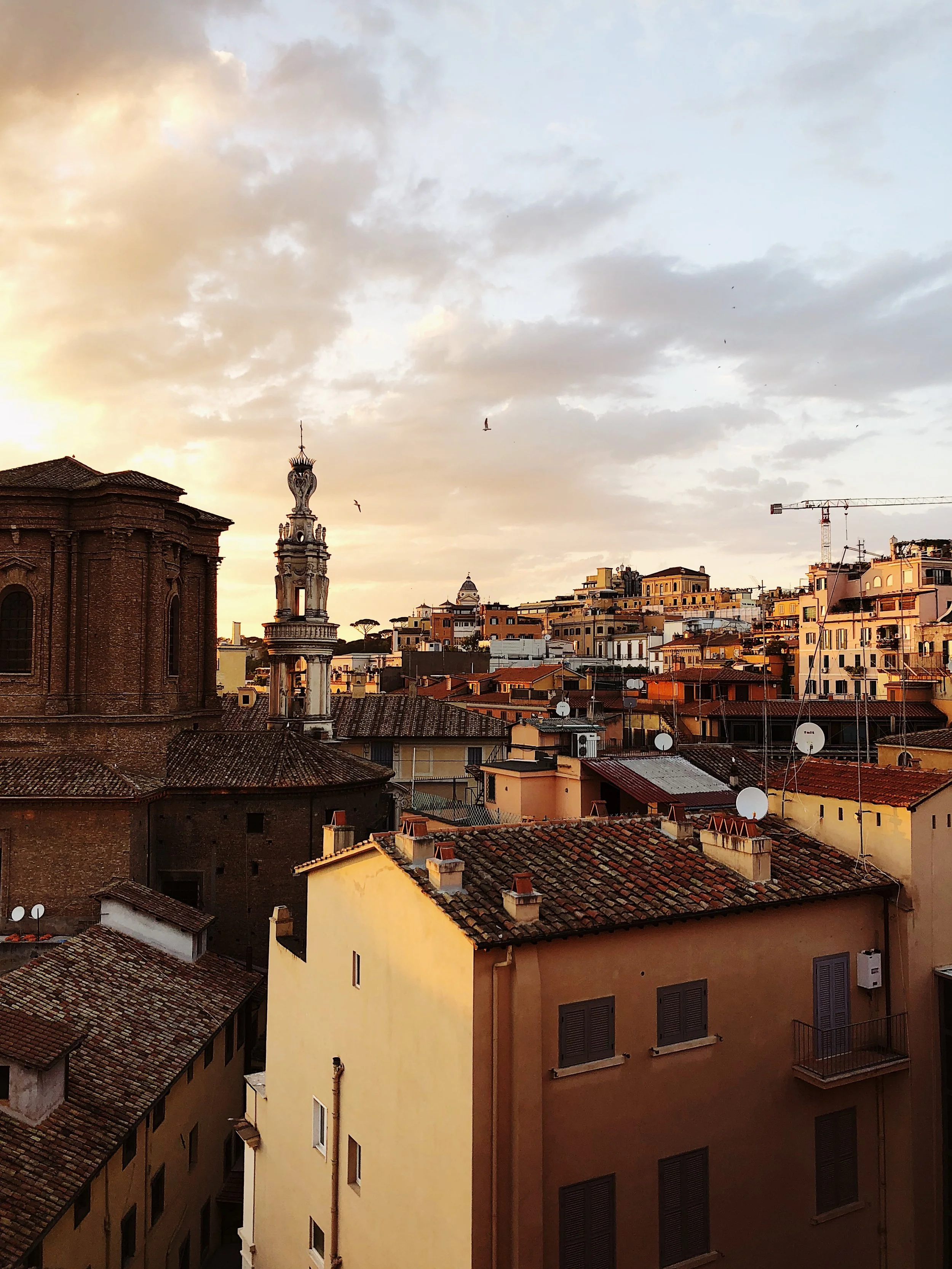 Sunset view over a city with terracotta rooftops, historic buildings, and a tall ornate tower, under a partly cloudy sky.