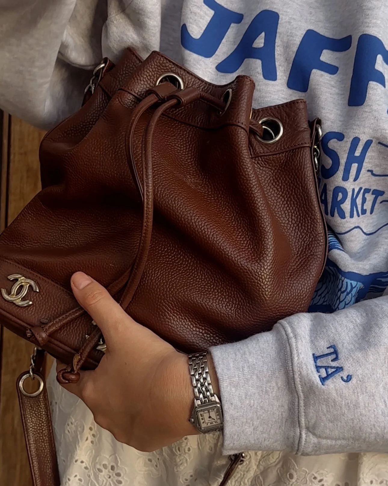 Person holding a brown leather handbag with a silver logo and eyelets, wearing a gray sweatshirt with blue text and a silver watch.