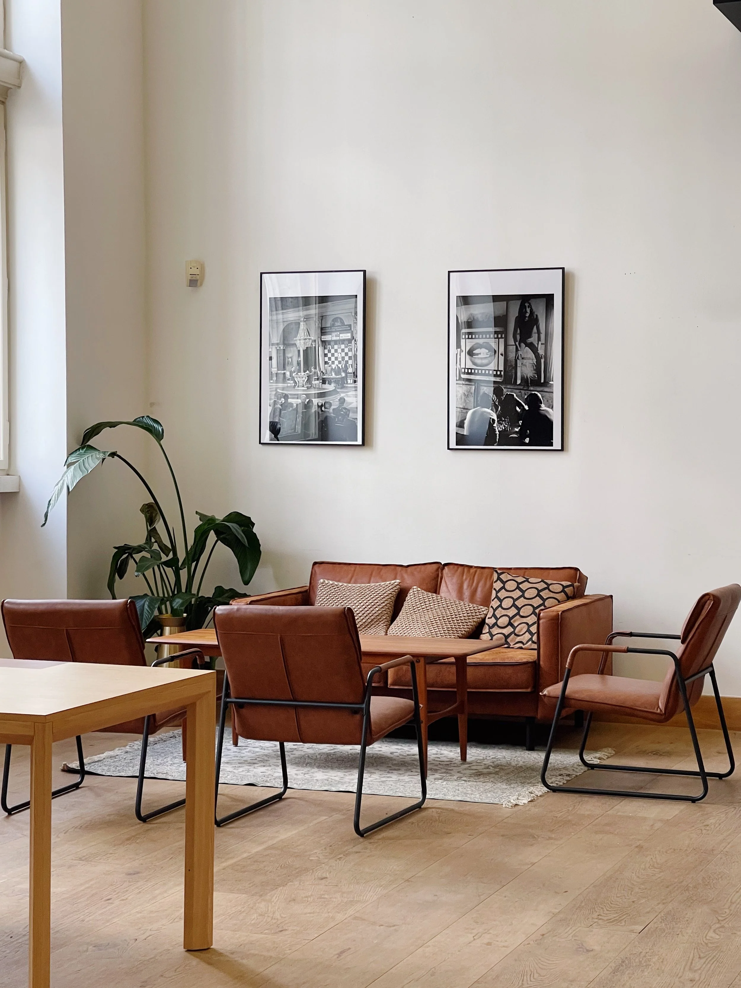 Living room with a brown leather sofa, a matching armchair, and two additional chairs with metal legs and pinkish upholstery. A small wooden table is in front of the sofa. There are two black-and-white framed photographs on the off-white wall above t