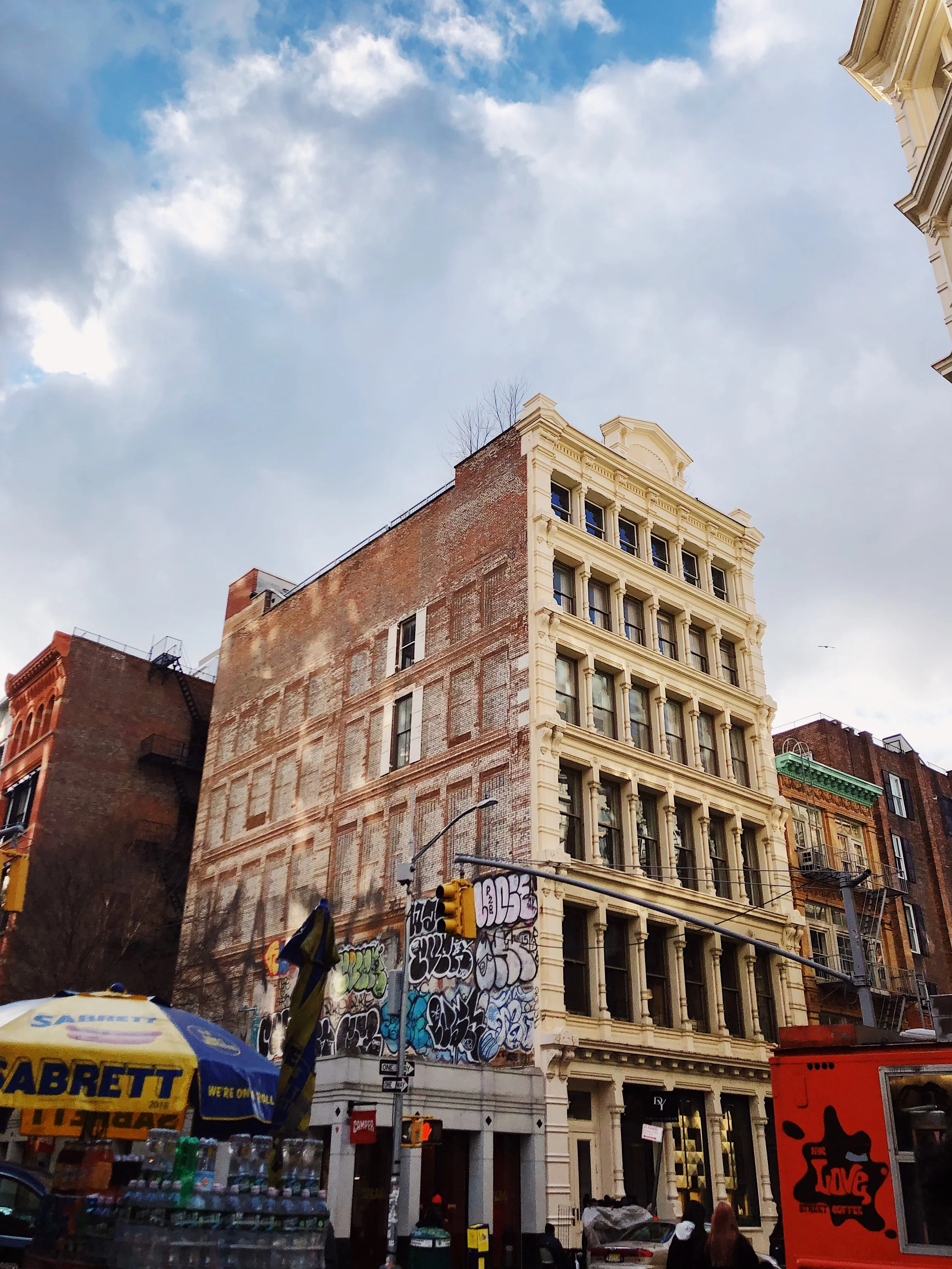 City street scene featuring a multi-story building with ornate details, graffiti on the lower wall, storefronts, and a yellow umbrella with 