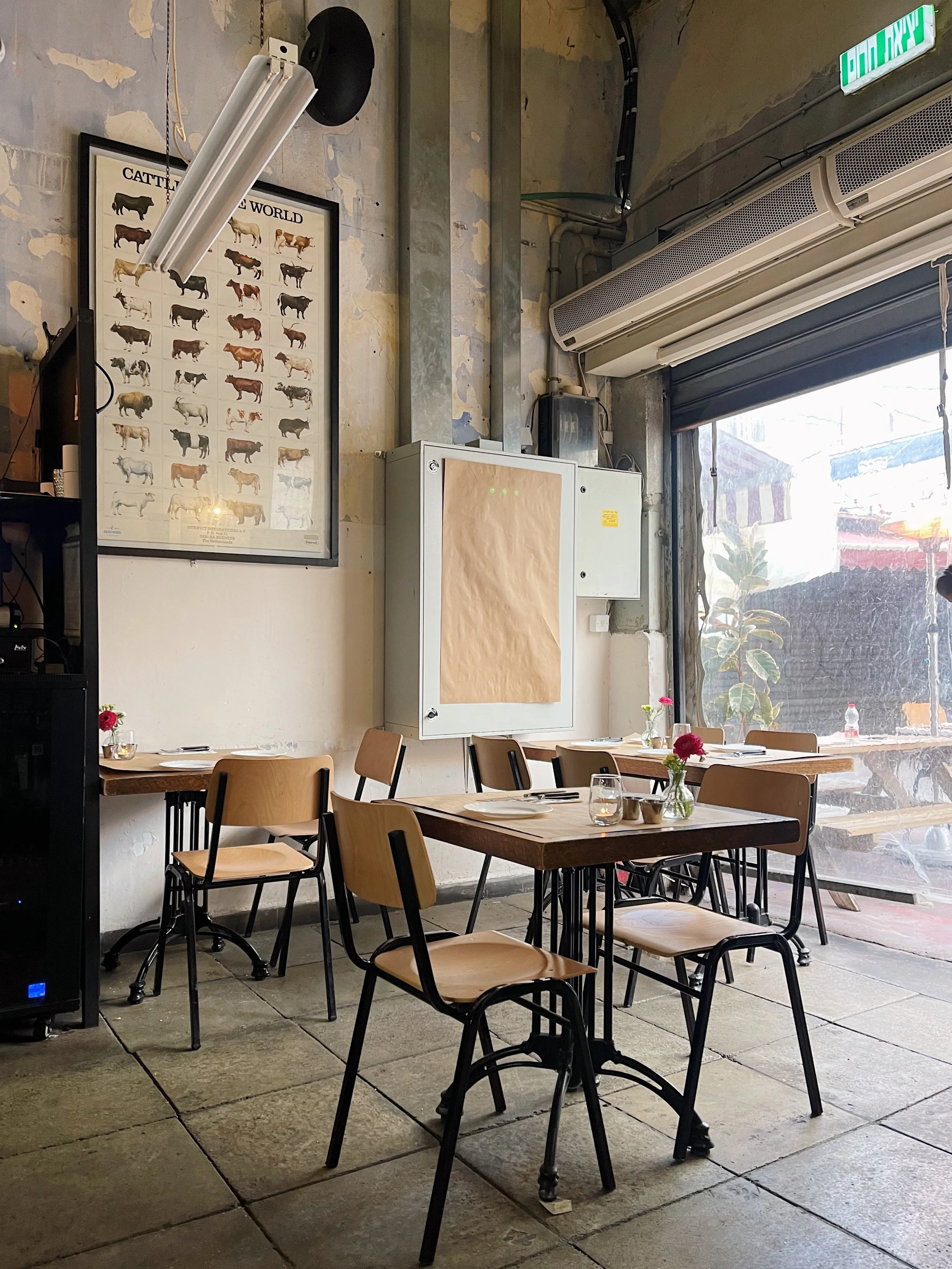 Empty restaurant with wooden tables and chairs, a wall-mounted poster of cattle breeds, and a large window with an outdoor view.