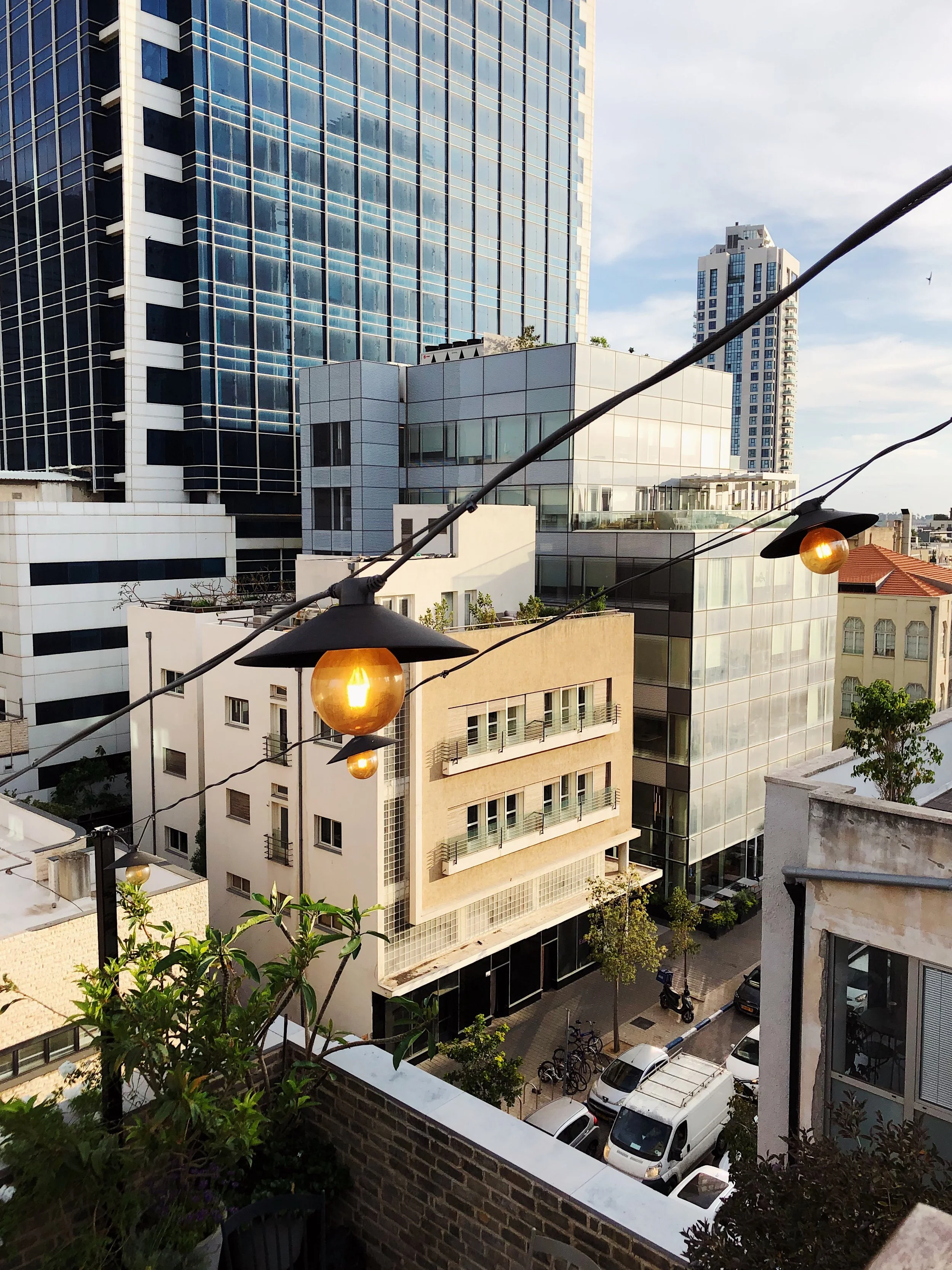 Urban cityscape featuring modern glass buildings and a street with parked cars beneath string lights.