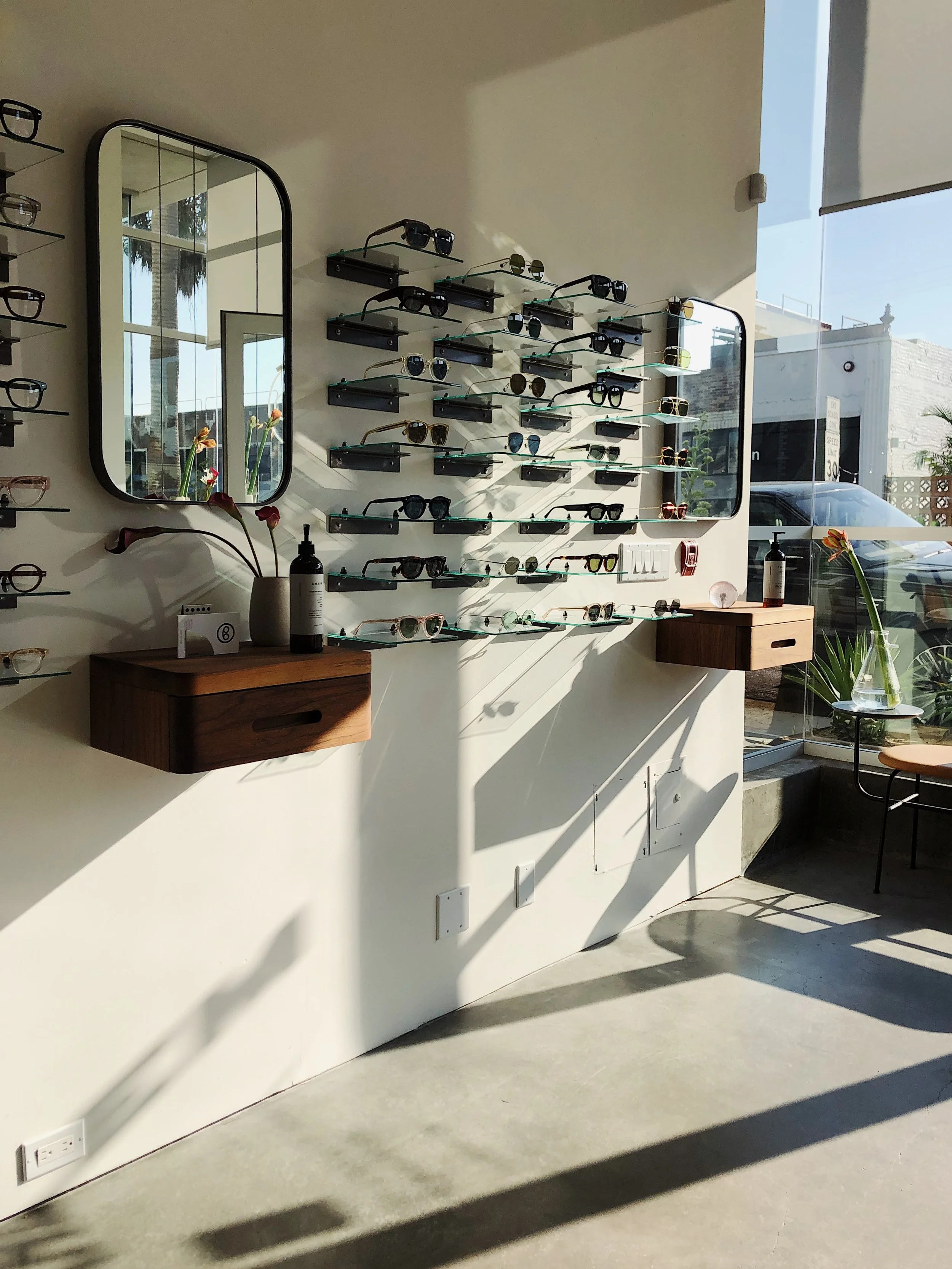 Display of various sunglasses on glass shelves with mirrors and wooden tables in a well-lit store, sunlight streaming through large windows.