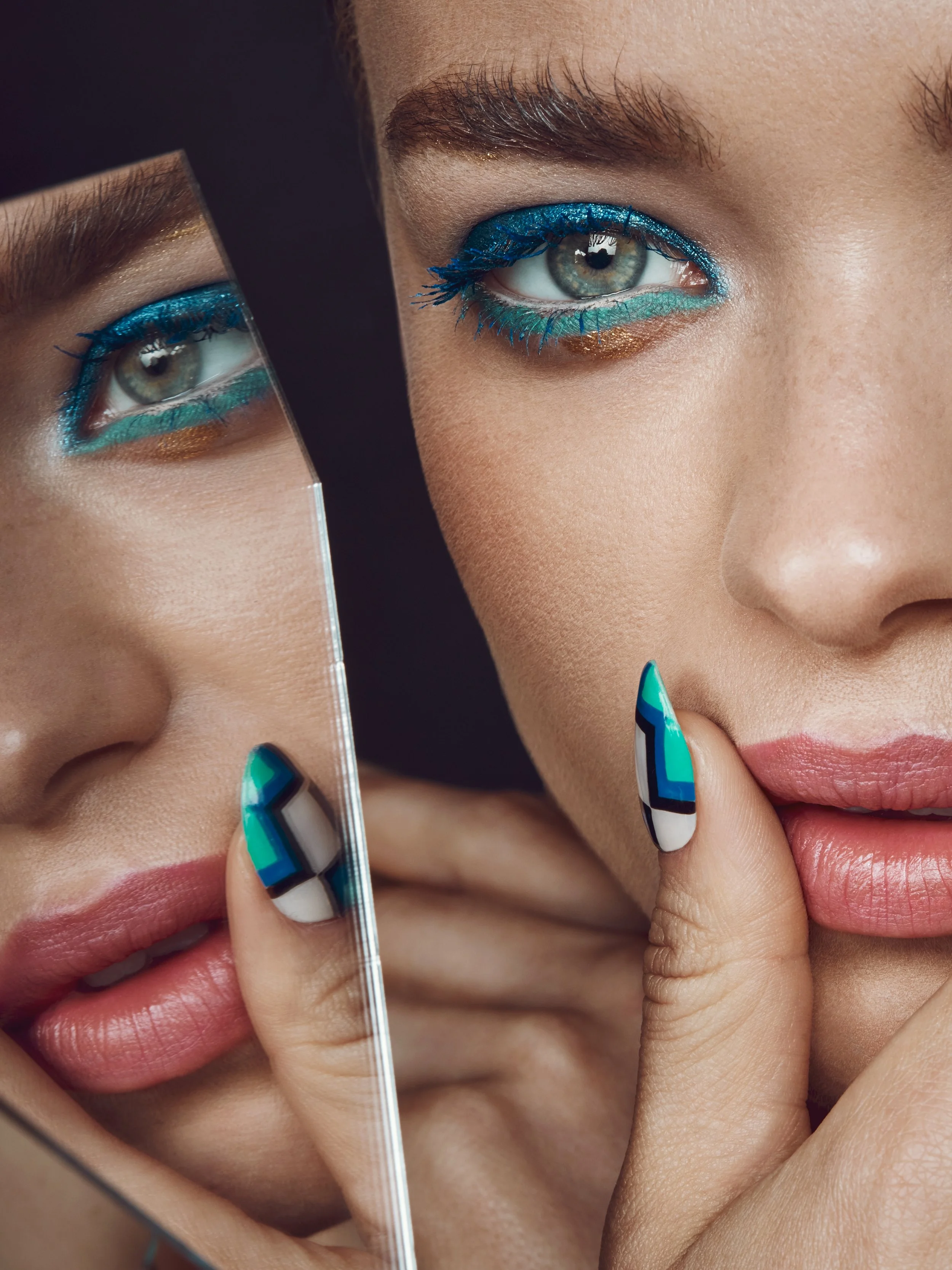 Close-up of a woman's face with colorful makeup, blue eyeshadow, and patterned nails, holding a mirror reflecting her lips and eye.