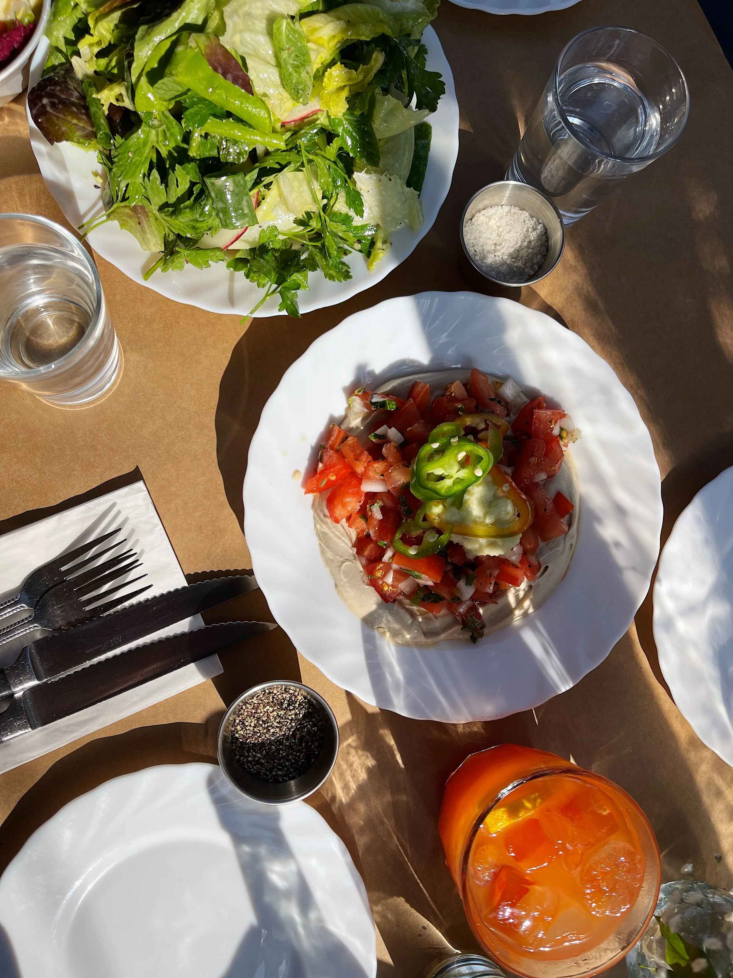 A table with a salad, a plate of tostada with tomatoes and jalapenos, glasses of water with ice, and condiments including ground black pepper and grated cheese.