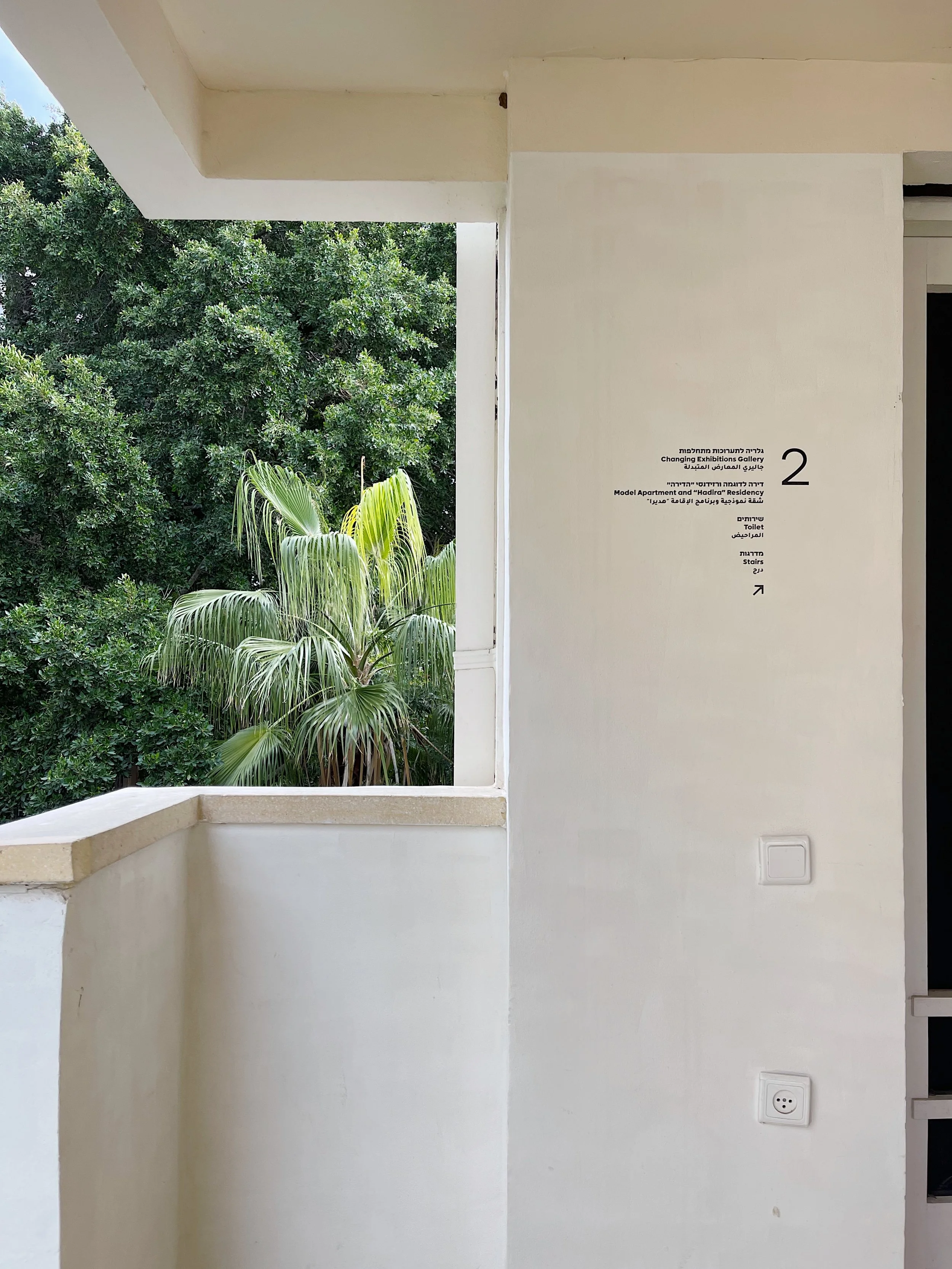 Interior view of a balcony with a white wall, sign indicating the location of changing exhibitions gallery, model apartment, and staircase, with lush green trees visible through the open window.