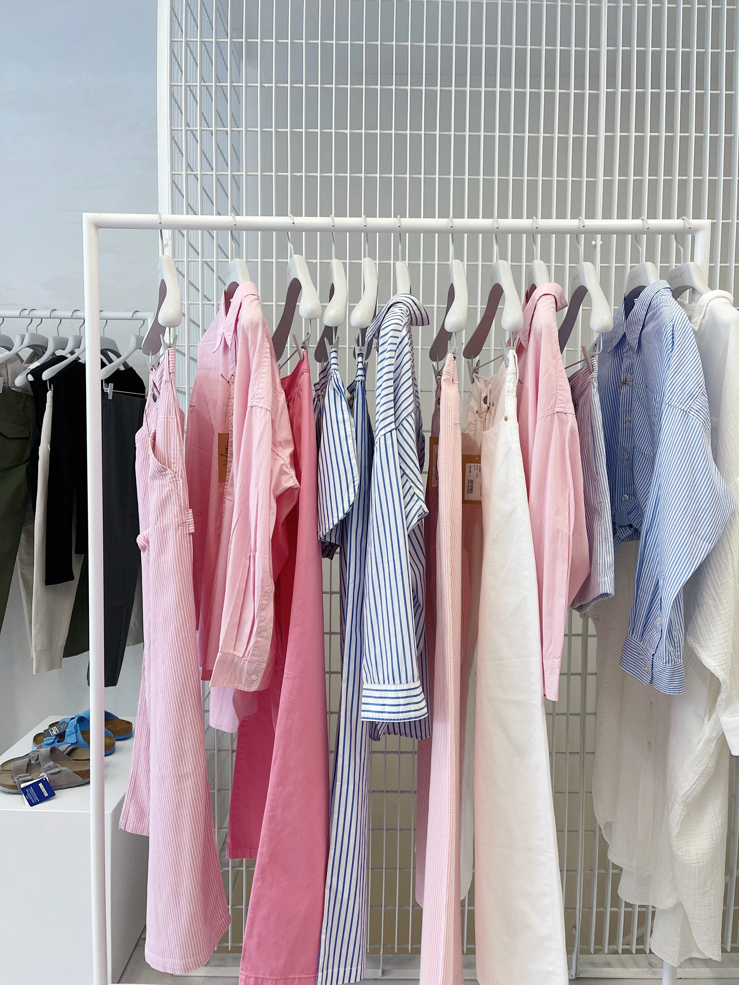 Clothing rack with various long-sleeved shirts and pants in pink, blue, white, and striped patterns, displayed on hangers inside a store fitting room.