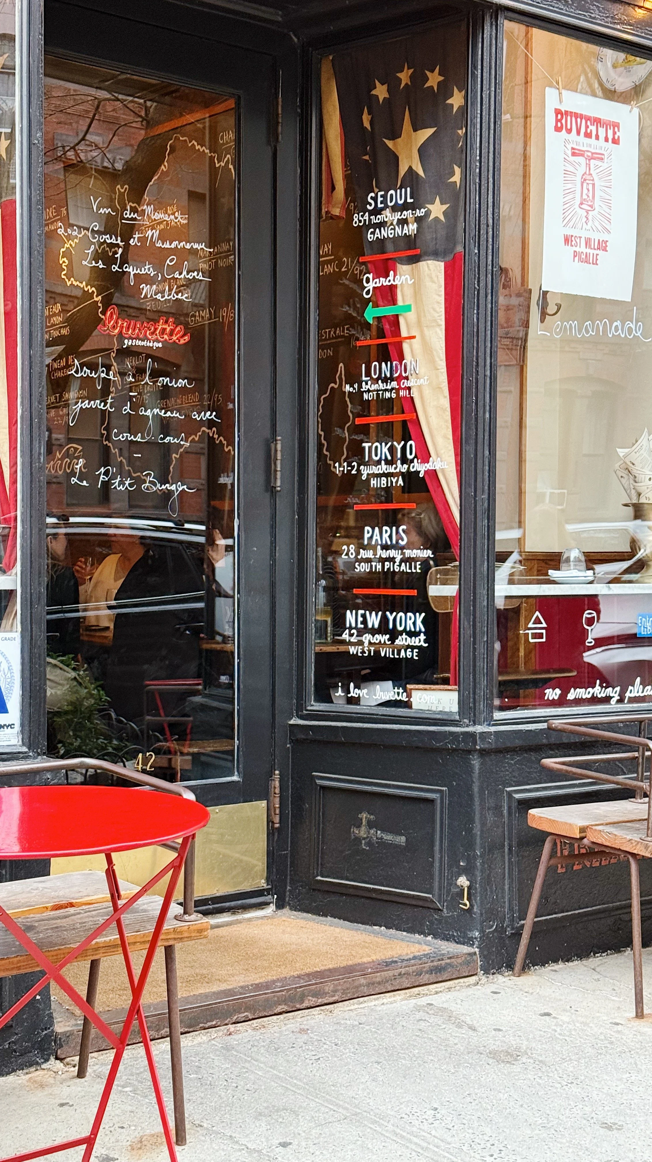 A restaurant window with a list of cities and addresses, a decorative flag with stars, and an illustrated map of Europe and France. There is a red table outside and a sign that says 'Buvette West Village Pigalle.'