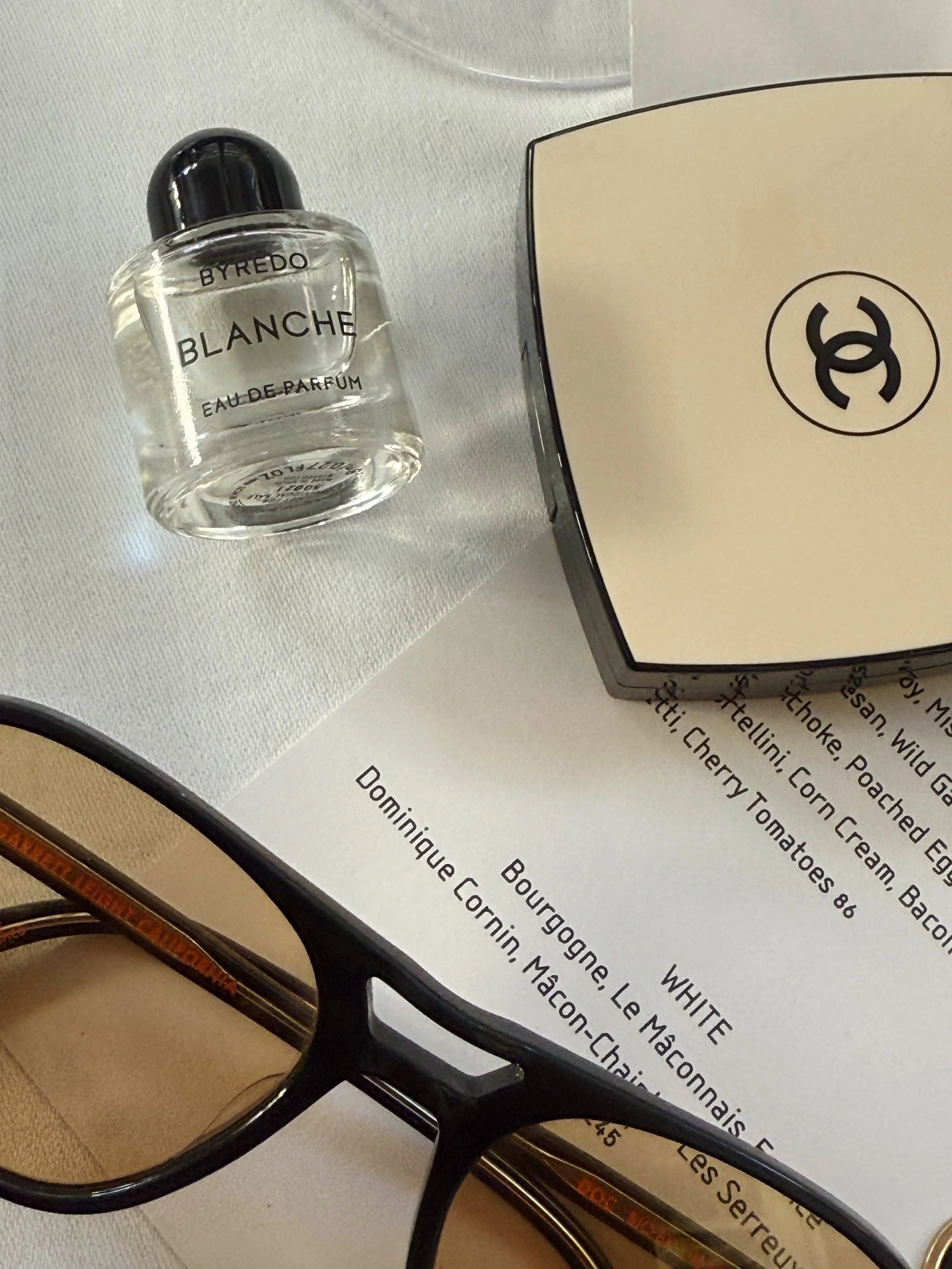 A bottle of Byredo Blanche Eau de Parfum, a Chanel compact, and black eyeglasses on a table with a printed menu and a white cloth.