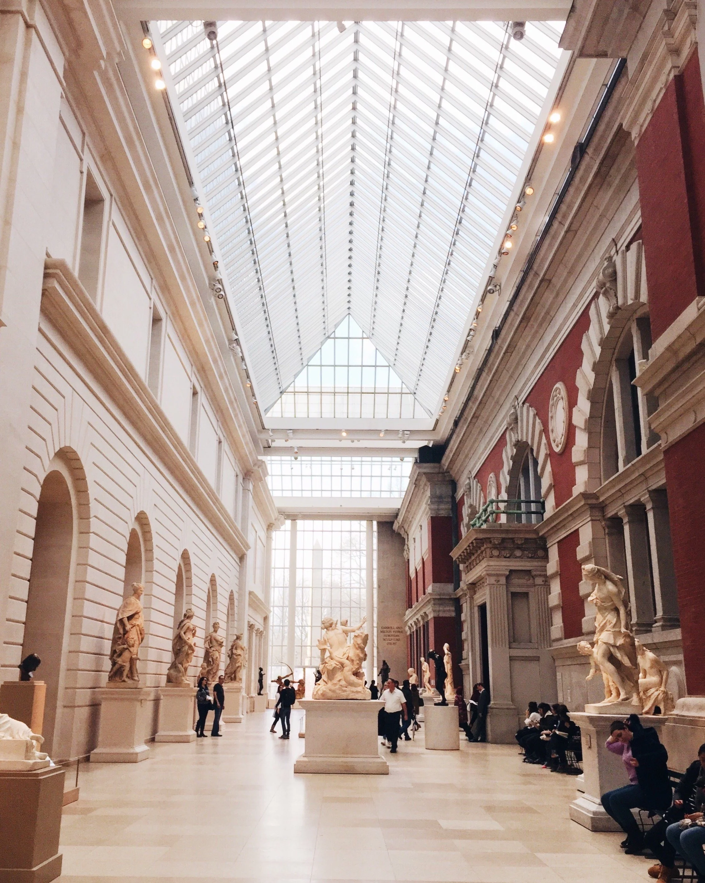 Interior view of a museum hall with classical sculptures displayed on pedestals, large windows, and a glass ceiling allowing natural light.