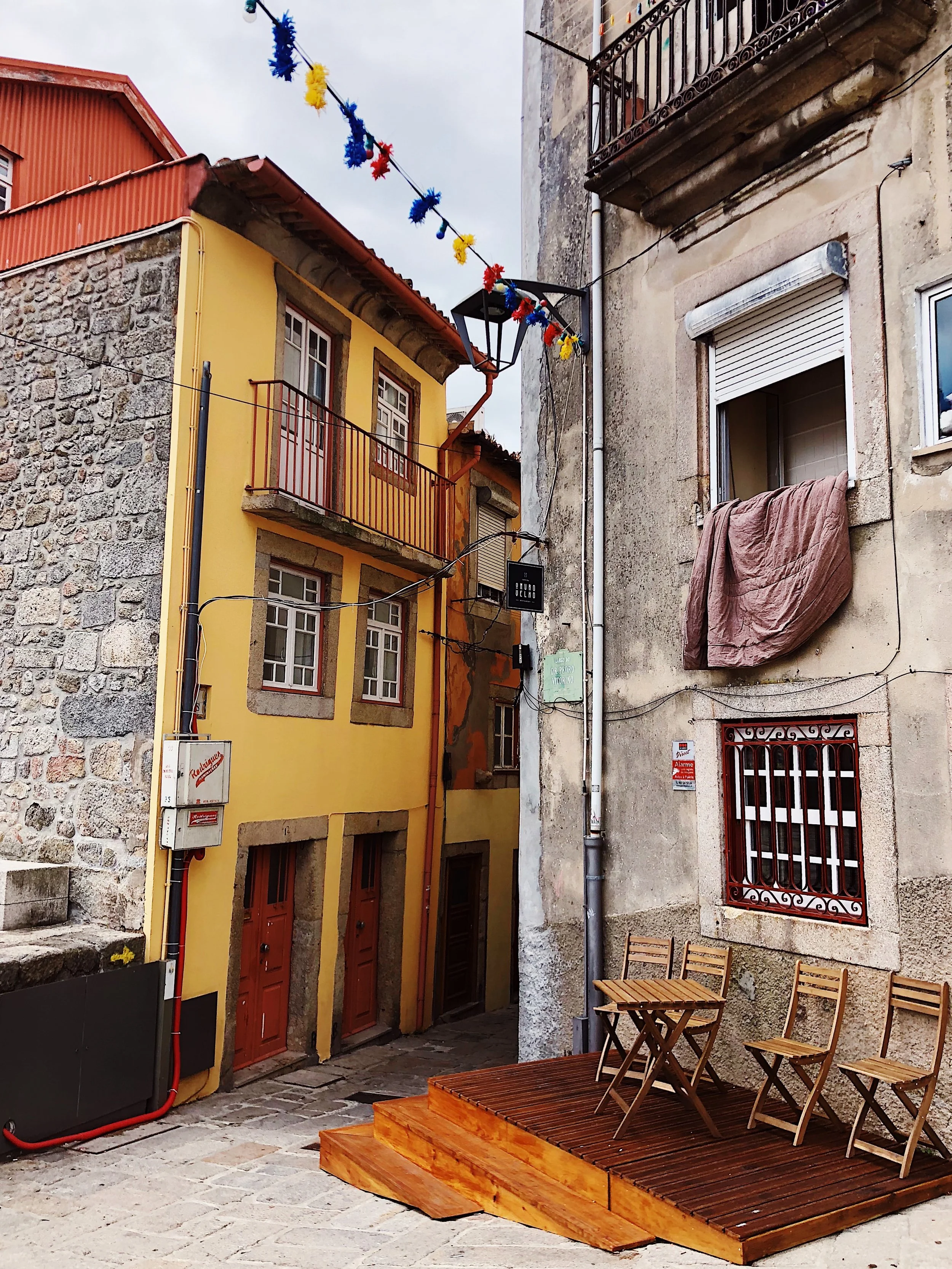 A narrow alley with colorful buildings, including a yellow and a gray building, and a small wooden deck with chairs outside, overhead string decorations, and a streetlamp.