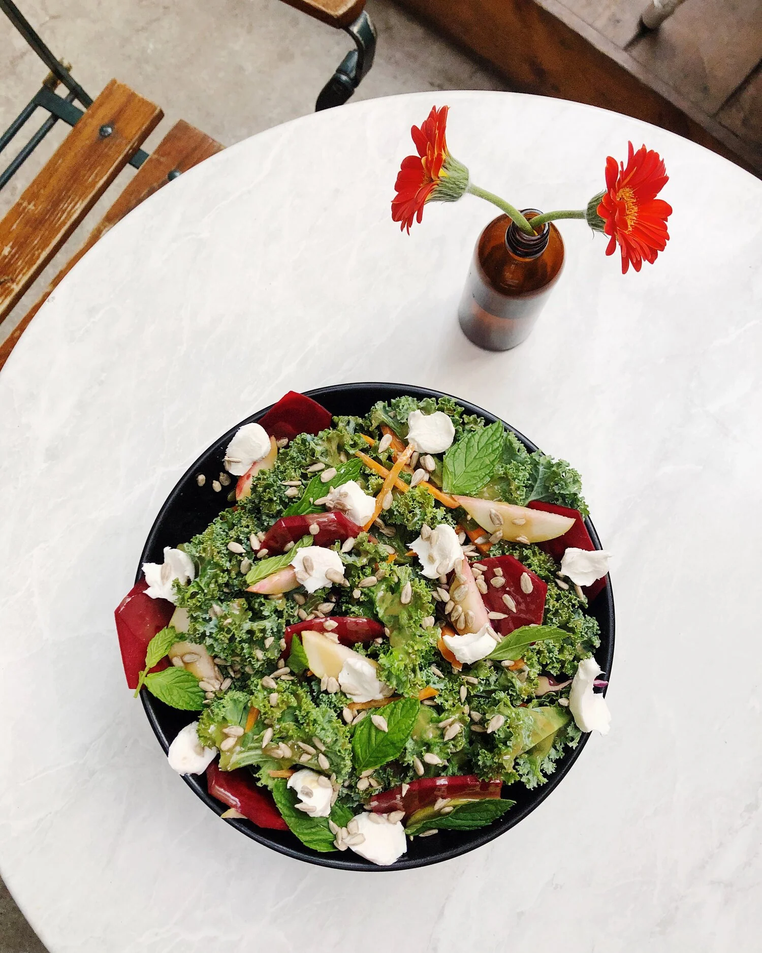 A bowl of mixed greens salad with vegetables, cheese, sunflower seeds, and herbs, on a white marble table. A small brown vase with orange-red flowers is in the background.