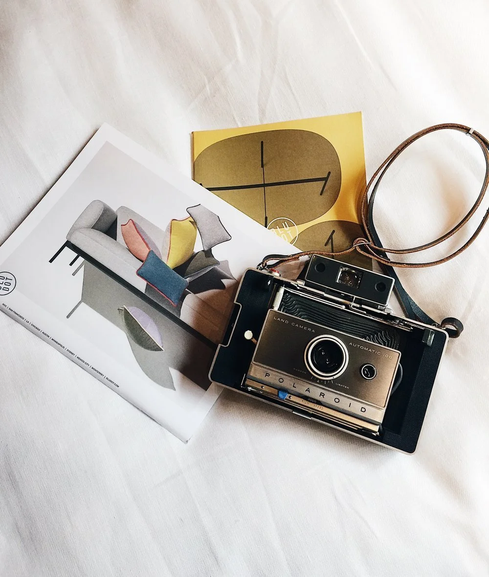 A vintage Polaroid camera, a yellow clock card, a piece of artwork, and a magazine with a modern sofa on the cover are arranged on a white surface.