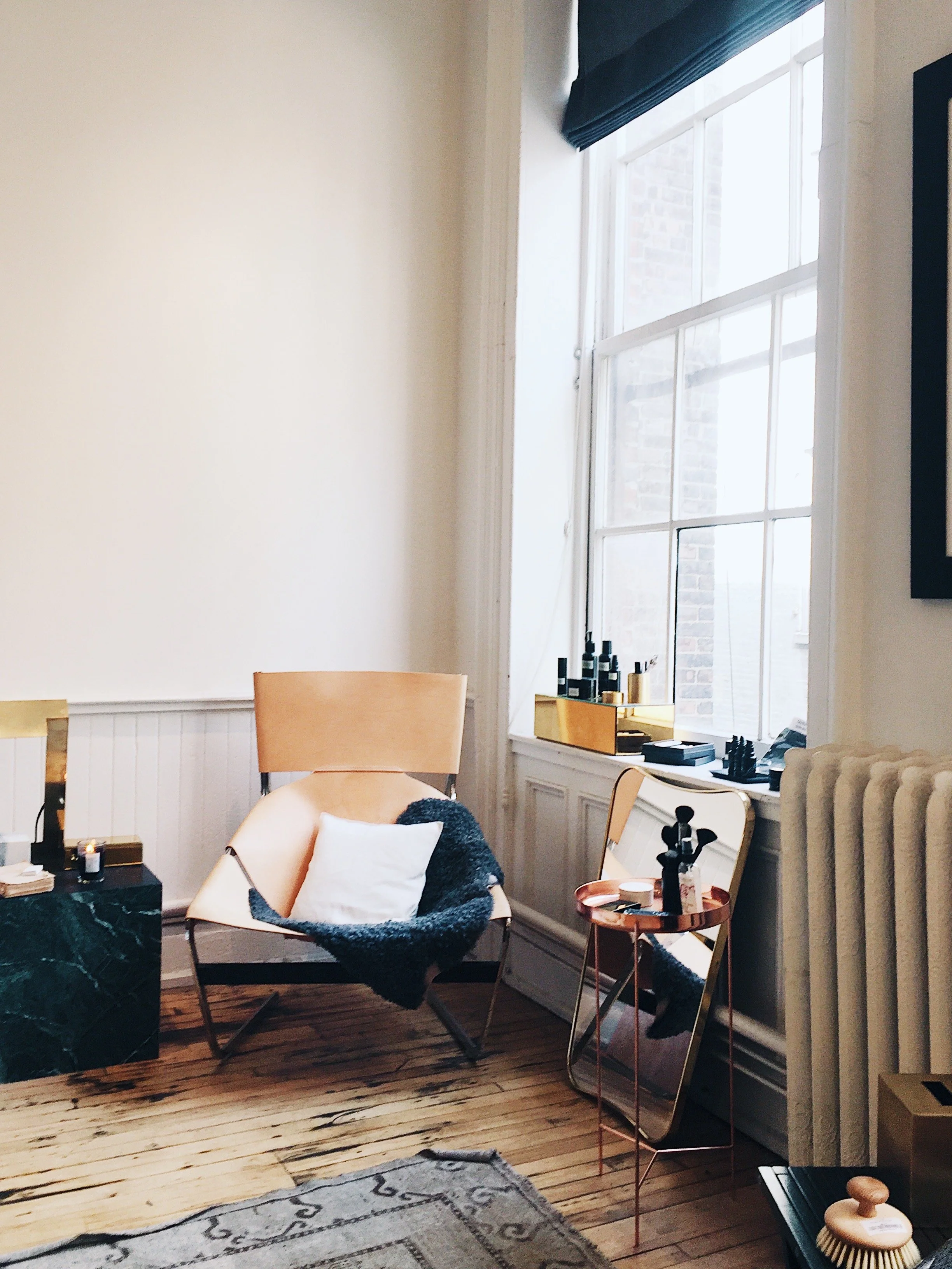 A cozy corner in a room with a large window, a tan modern armchair with a white pillow and a black throw, a small copper side table with skincare products, a mirror propped against the wall, and a wooden floor with a patterned rug.