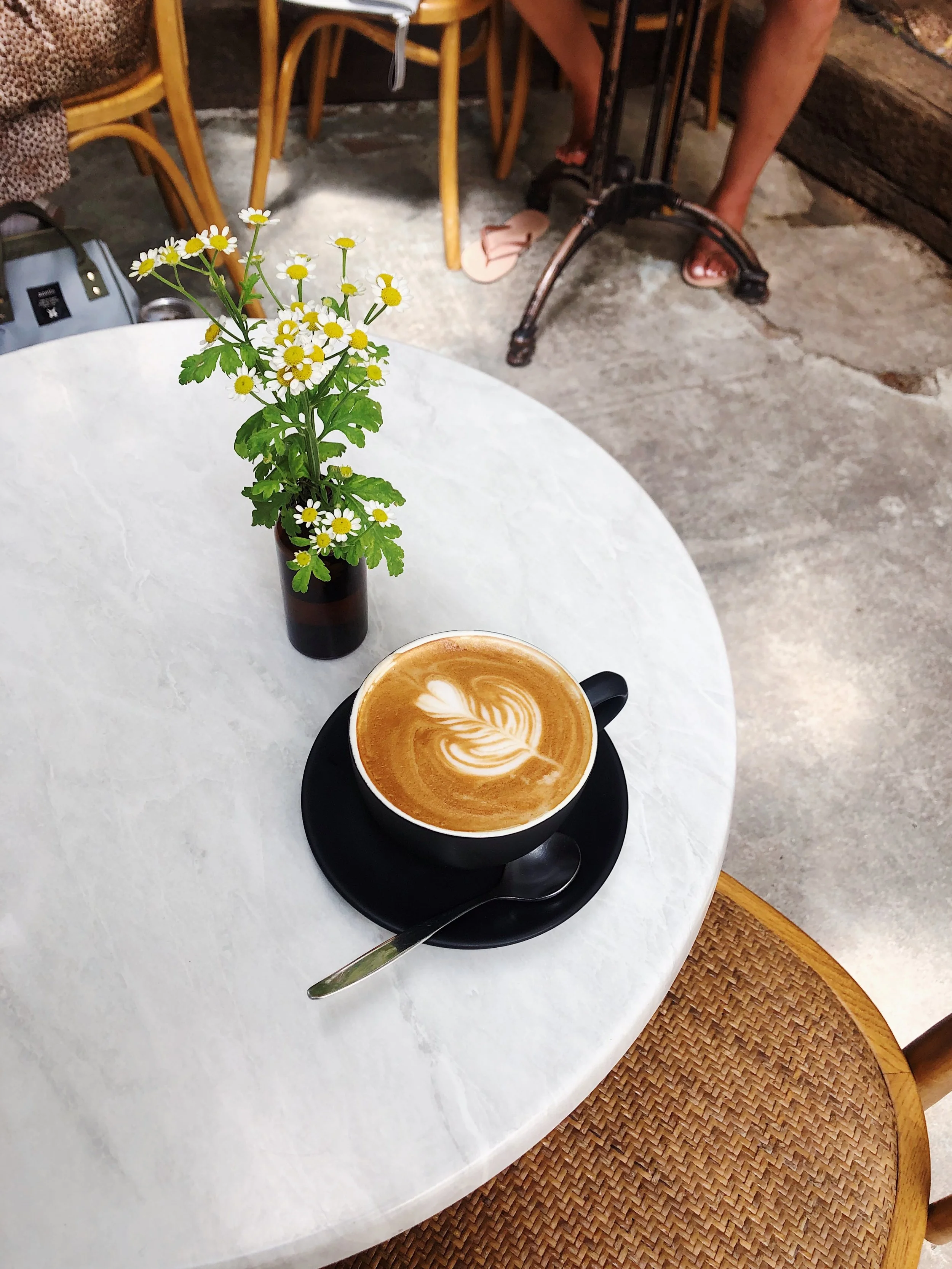 A cup of latte with latte art on a black plate with a silver spoon, a small vase with white daisies, sitting on a white round table in a cafe with a chair and part of a person visible in the background.