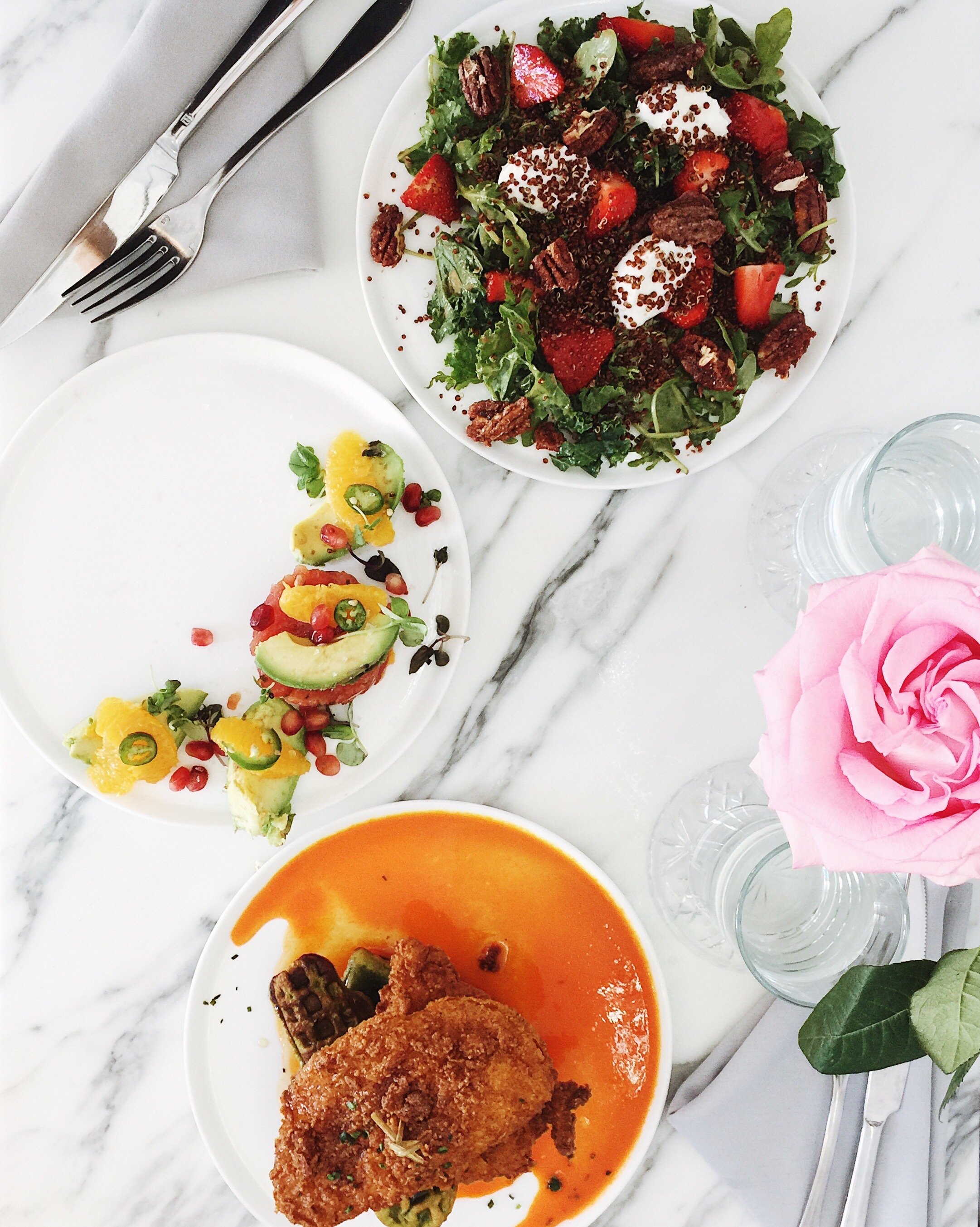 A top view of three plates of food including a salad with strawberries and chocolate shavings, a colorful vegetable and avocado plate, and a fried chicken dish with sauce, alongside a pink rose, glasses, and cutlery on a white marble table.