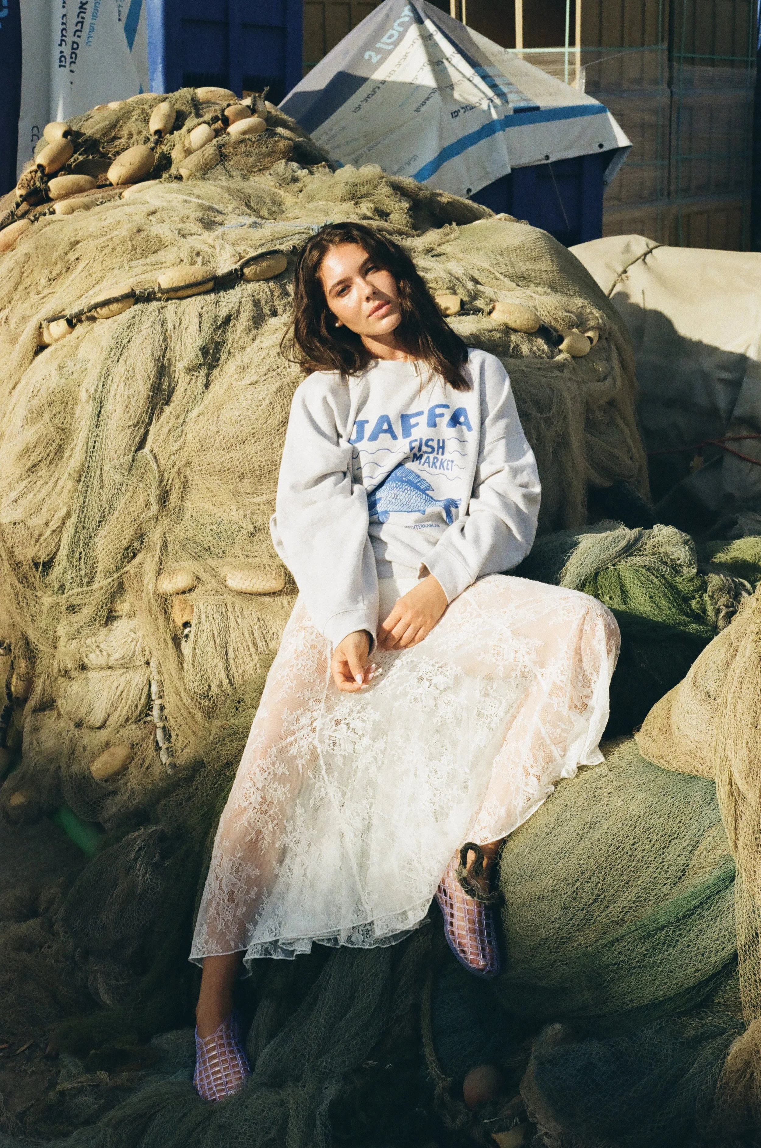 A woman with dark hair wearing a white graphic sweatshirt and a lace skirt, sitting on a pile of fishing nets and ropes at a fish market, with a blue and white canopy in the background.
