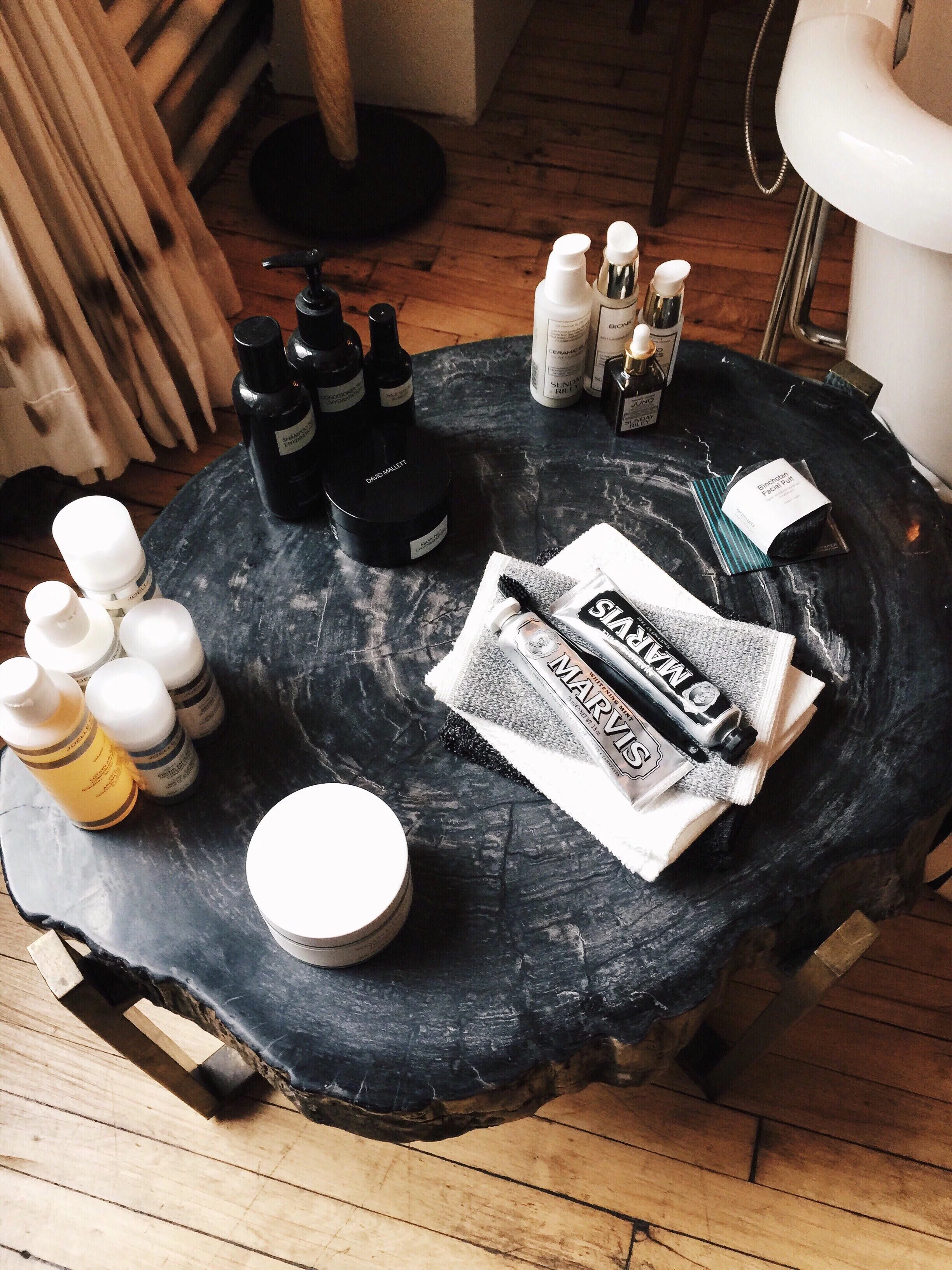 A black marble table with various skincare and grooming products, including bottles, jars, and tubes, arranged on it. Some items are placed on a cloth, and the table is positioned on a wooden floor with curtains and furniture visible in the backgroun