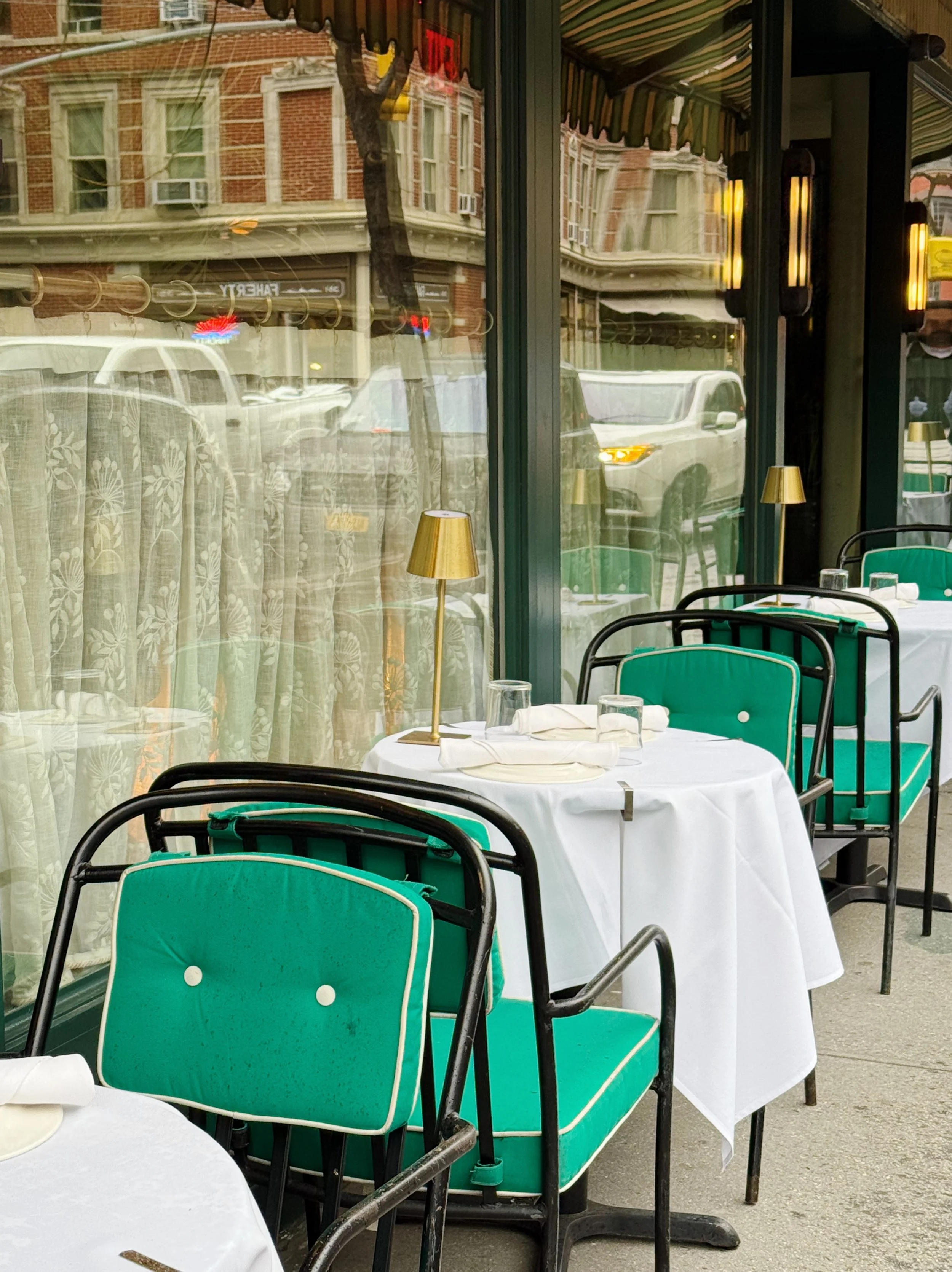 Empty outdoor restaurant tables with white tablecloths, green chairs, and gold-colored lamps, outside a storefront with large glass windows reflecting parked cars and buildings.