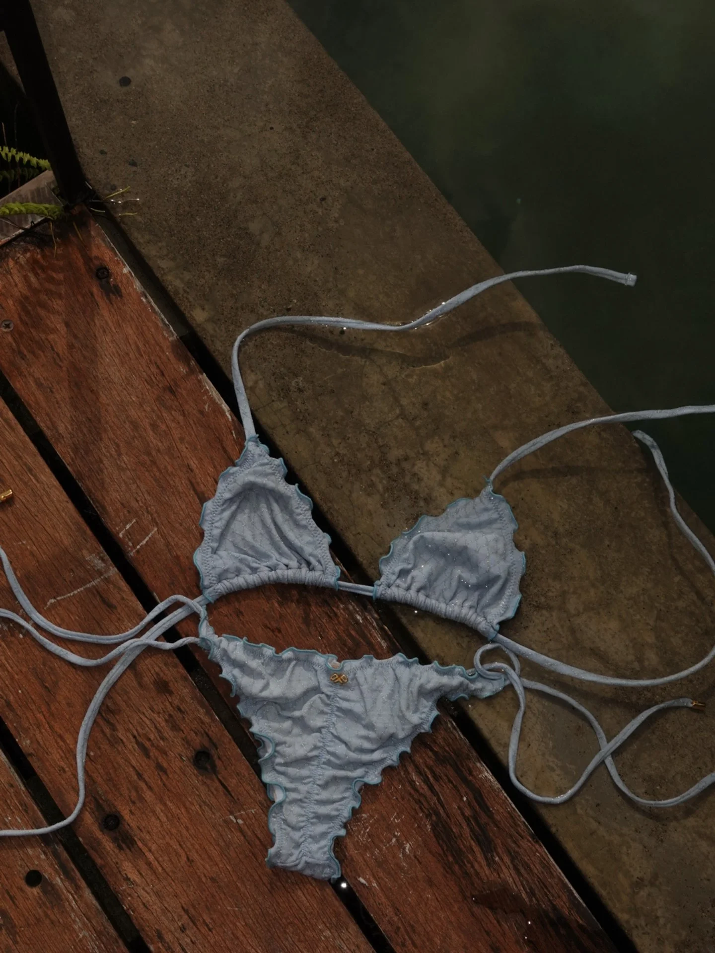 A light blue bikini with ruffled edges rests on a wooden dock next to water.