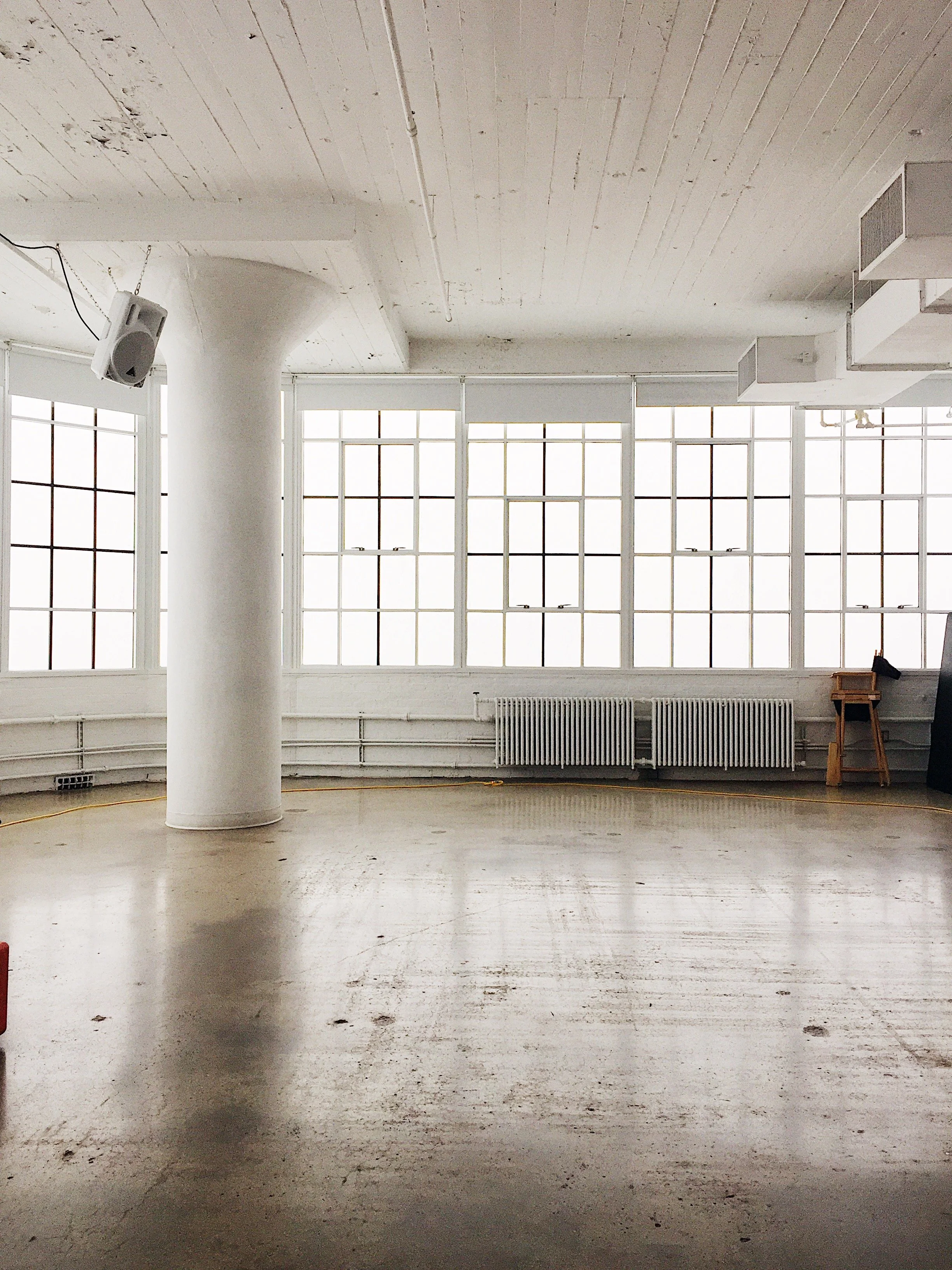 An empty room with large windows, white walls, and a concrete floor. There is a white pillar near the center, a radiator along the wall, a small wooden chair, and some equipment on the right side.