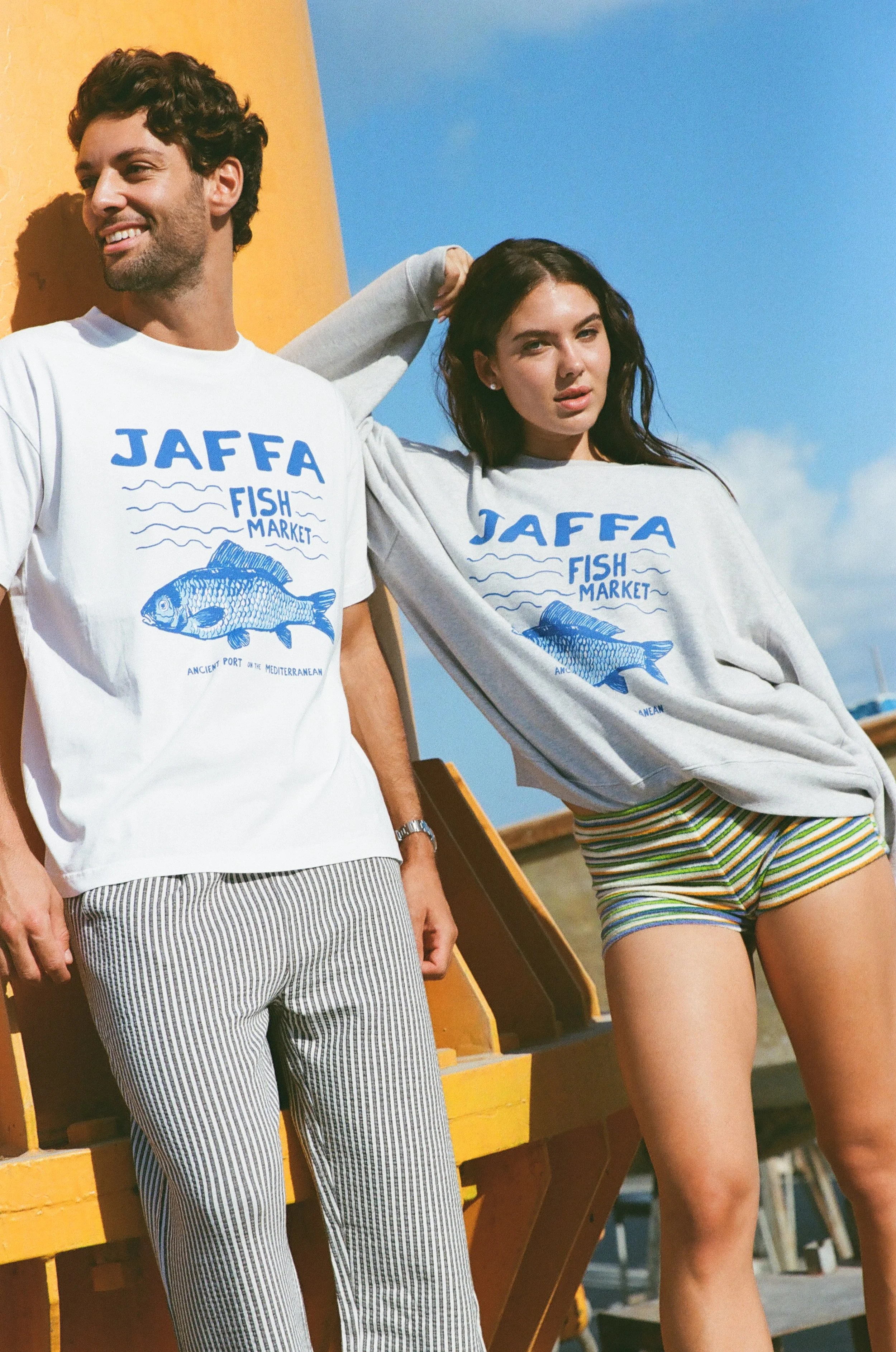 A man and a woman stand outdoors near a yellow and orange lifeguard tower, wearing matching Jaffa Fish Market T-shirts. The man has dark curly hair, a beard, and striped pants, while the woman has long dark hair and striped shorts. The sky is blue with some clouds in the background.