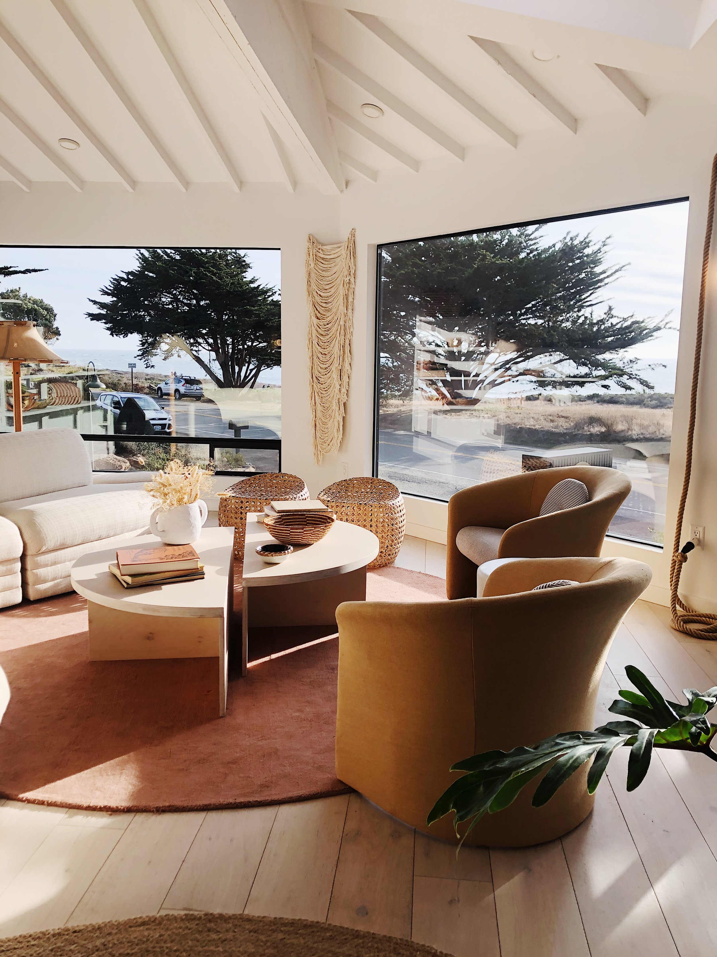 Cozy living room with large windows showcasing a view of trees and the outdoors, featuring a beige sofa, two tan armchairs, a white oval coffee table with decorative bowls and a book, and woven stool with a peach-colored rug.