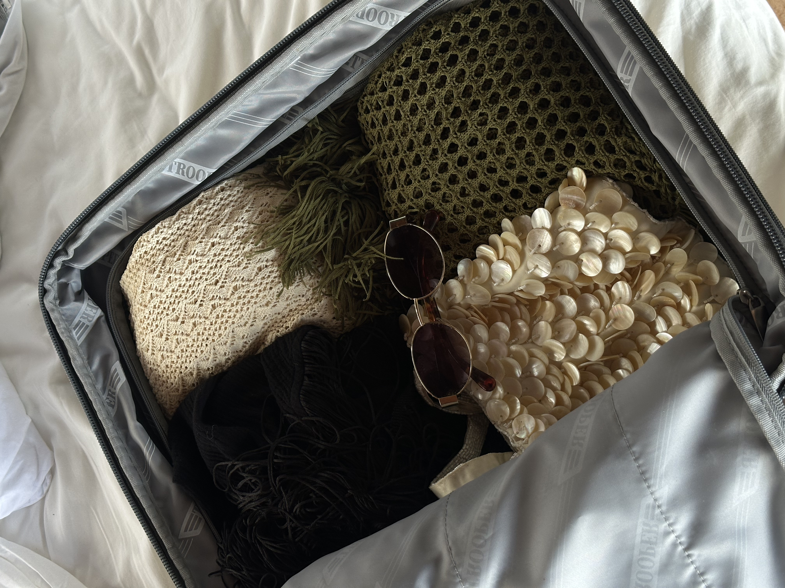 Open travel bag containing cream-colored shells, black string, beige netted fabric, green crochet fabric, sunglasses with dark lenses, and some greenery.