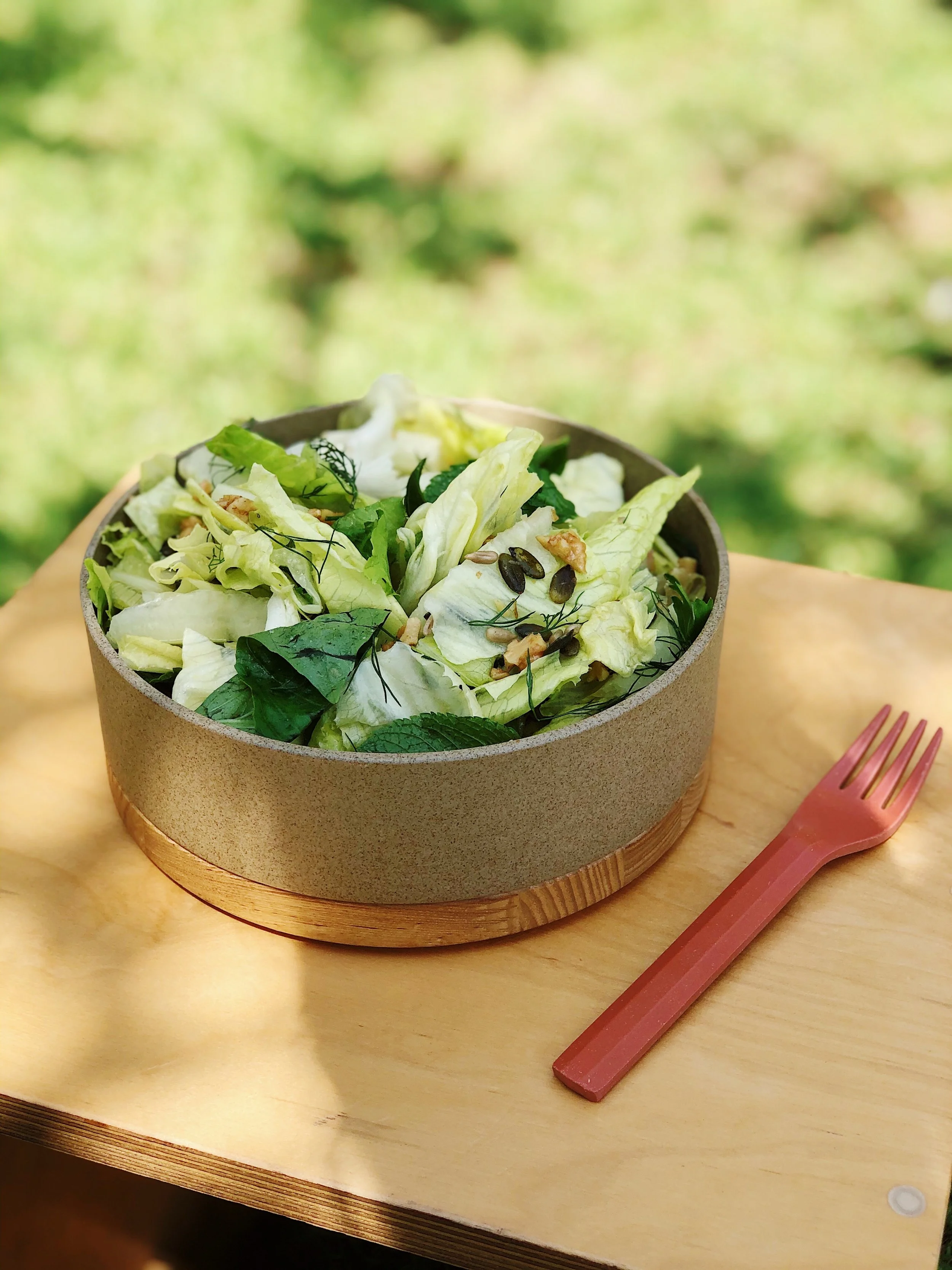 A bowl of mixed salad greens with seeds and herbs on a wooden table outdoors.