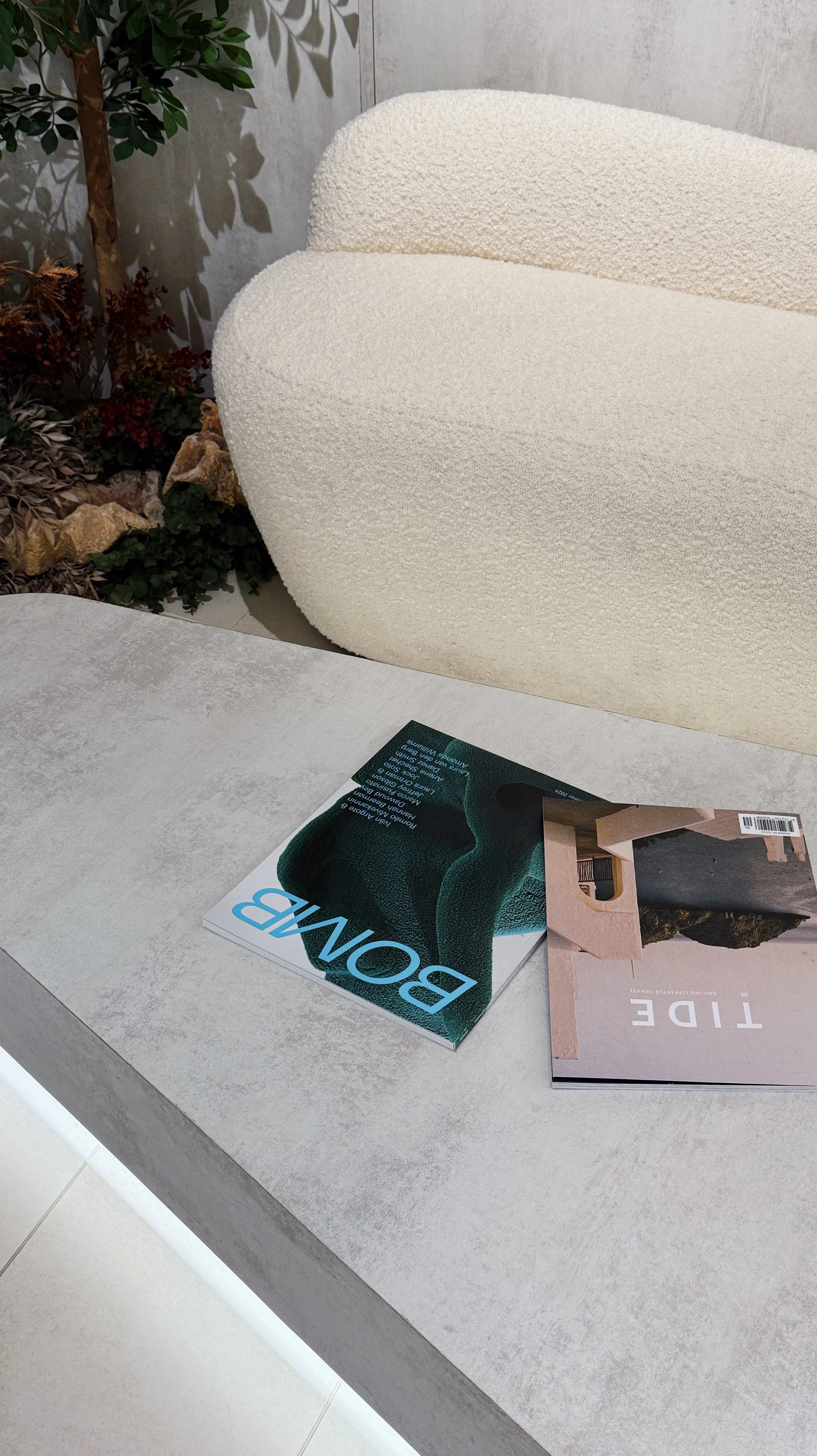 A magazine titled 'BONE' resting on a concrete table next to another magazine titled 'TIDE' in a modern interior setting with a white textured armchair and decorative plants.