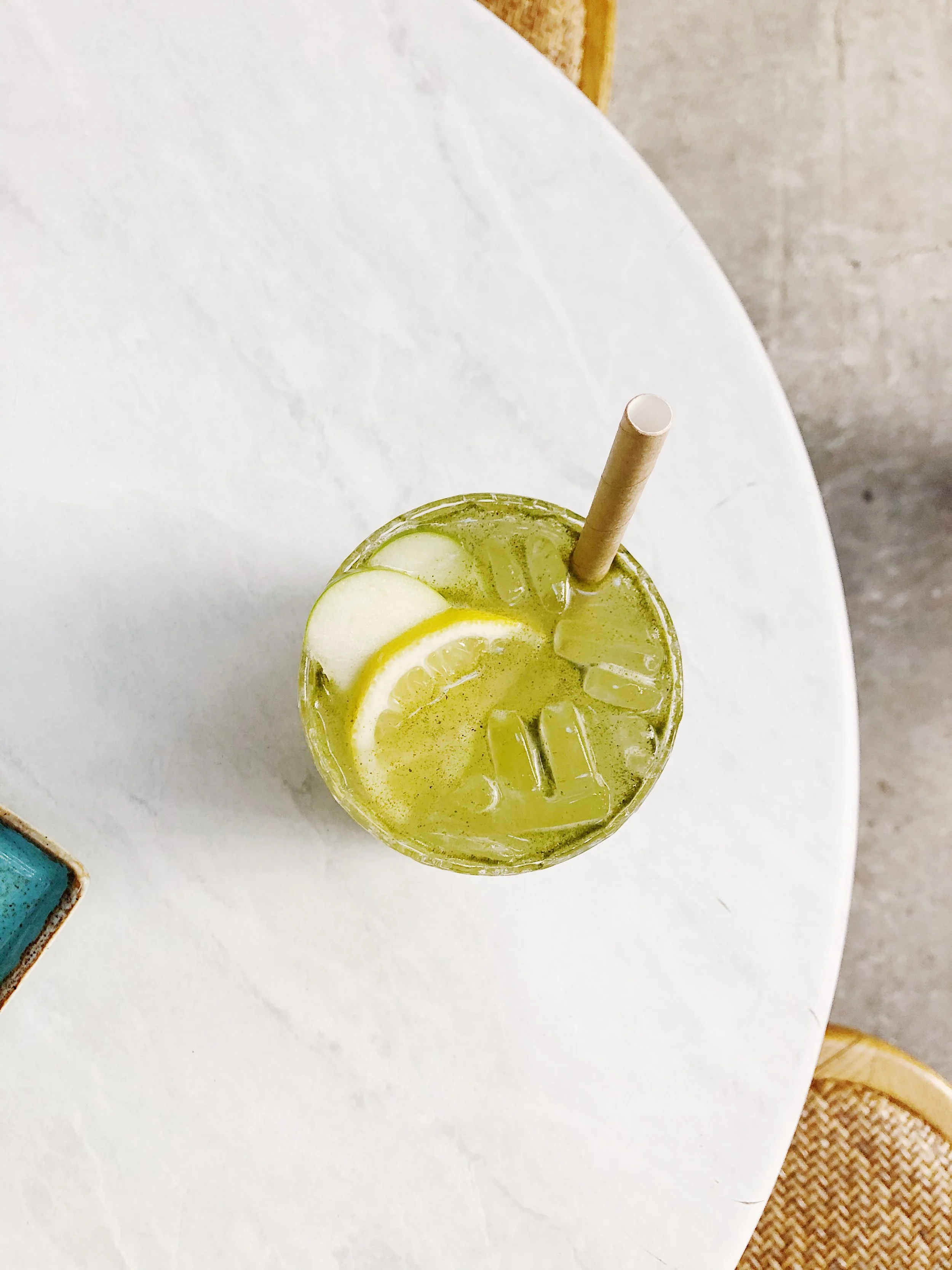Glass of lime and cucumber infused drink with ice cubes, served with a paper straw on a white table.