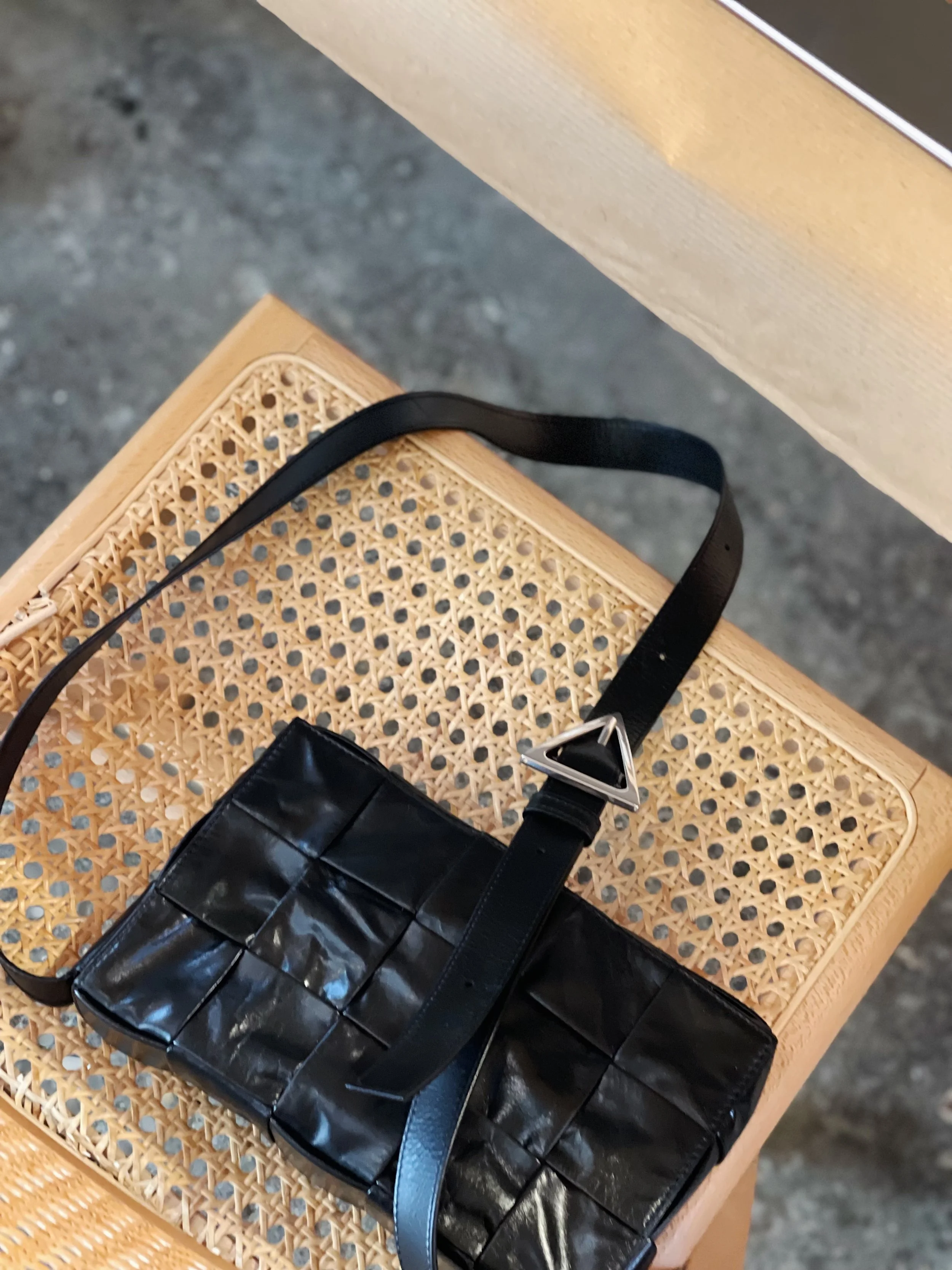 A black handbag with a woven pattern, resting on a wooden cane chair with a caned seat. The handbag has a long strap and a triangular metallic clasp.