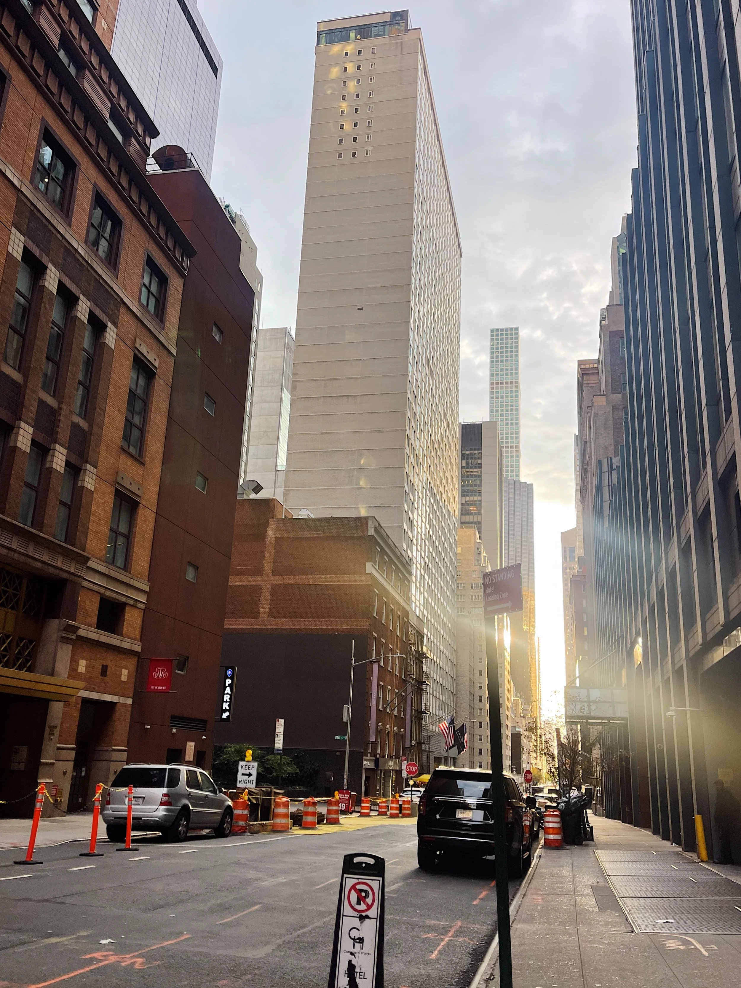 A city street scene at sunset with tall skyscrapers, parked cars, and construction cones, featuring a bright sun flare near the horizon.