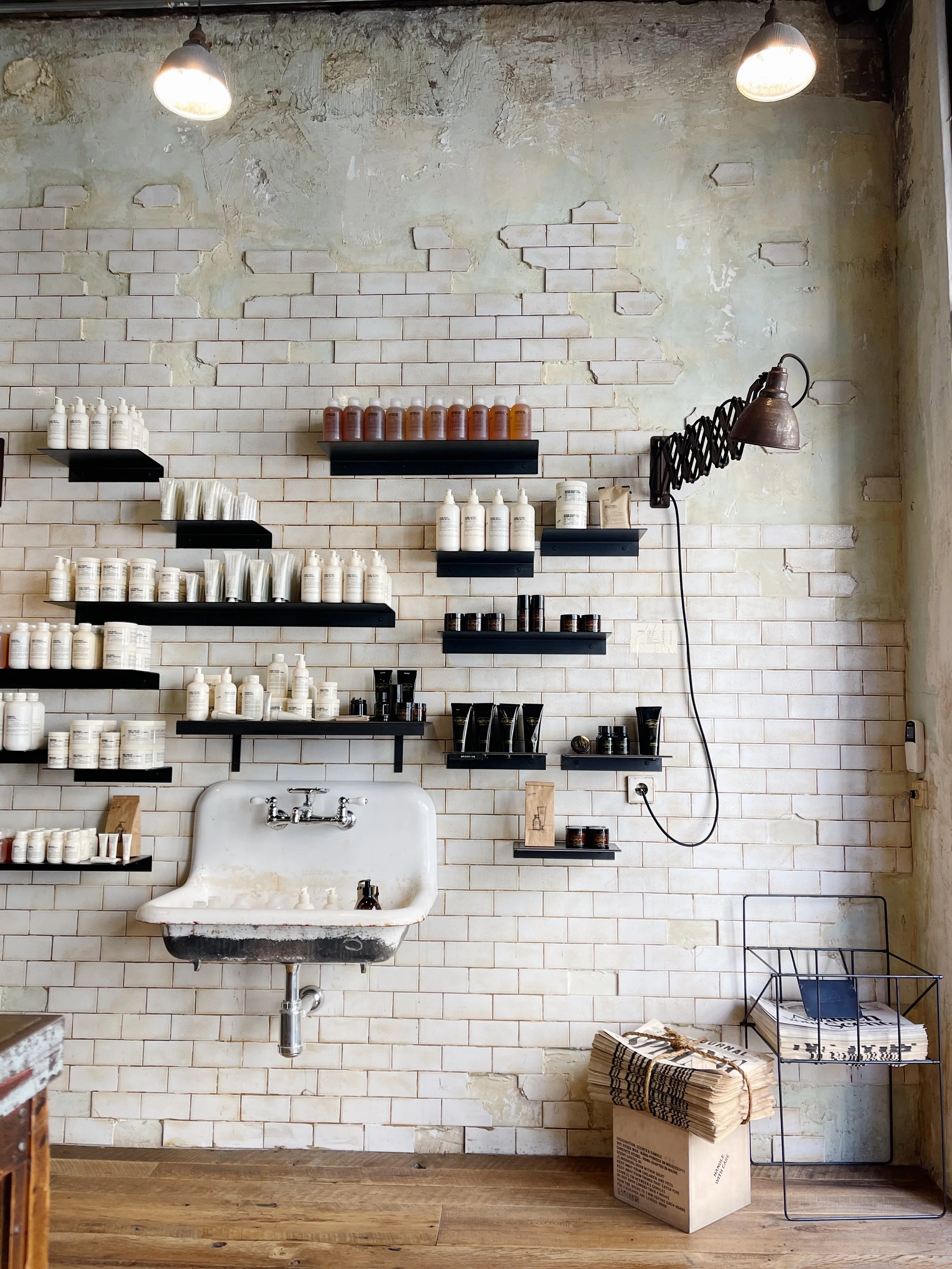 Interior of a rustic store with a vintage sink, shelves with bottles and jars, paper bundles, and a wire rack with newspapers. Exposed brick wall, wood floor, and hanging lamps.