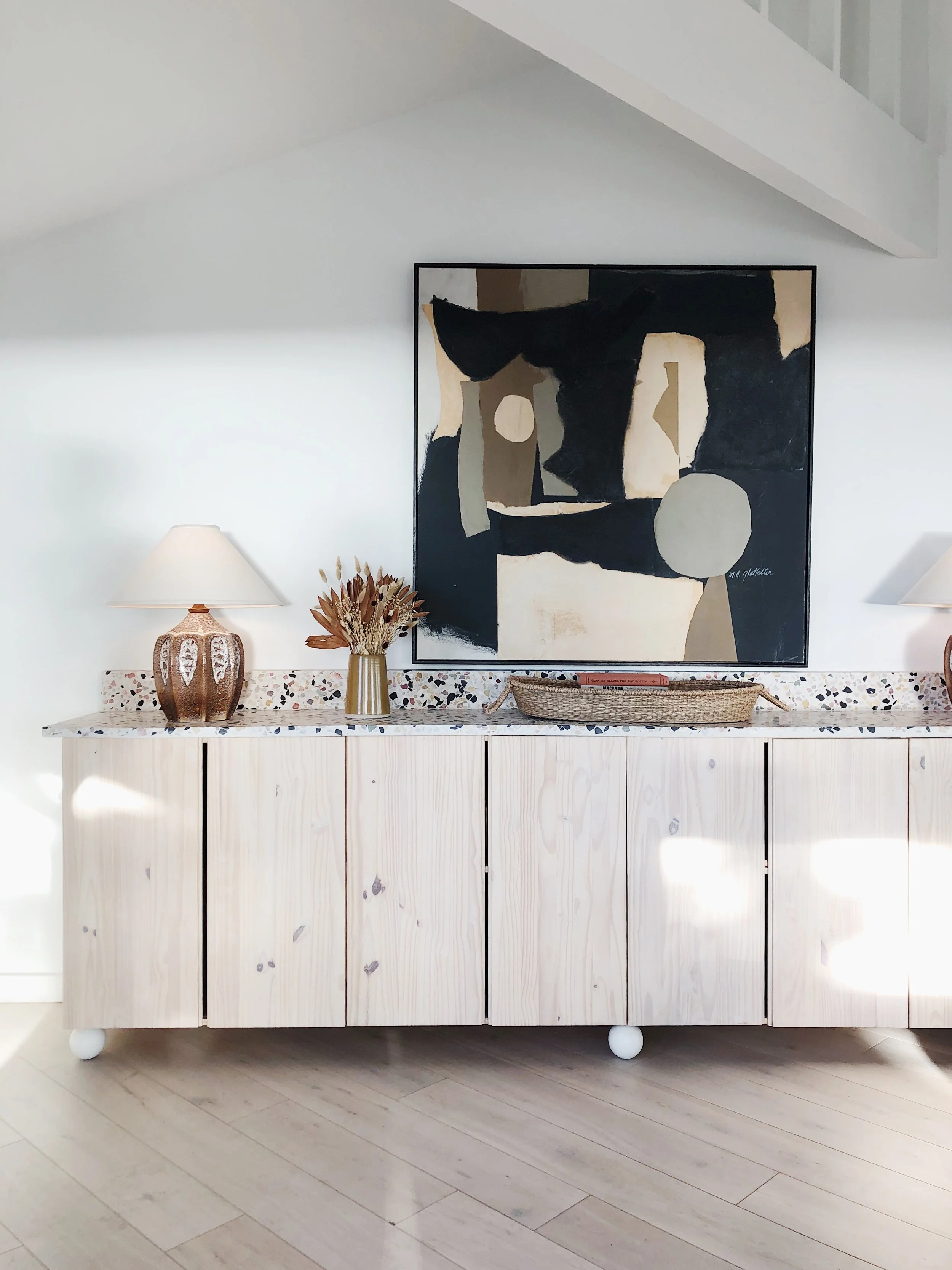 Decorative sideboard with an abstract painting, two table lamps, a vase with dried flowers, and a woven tray in a modern, minimalist room.