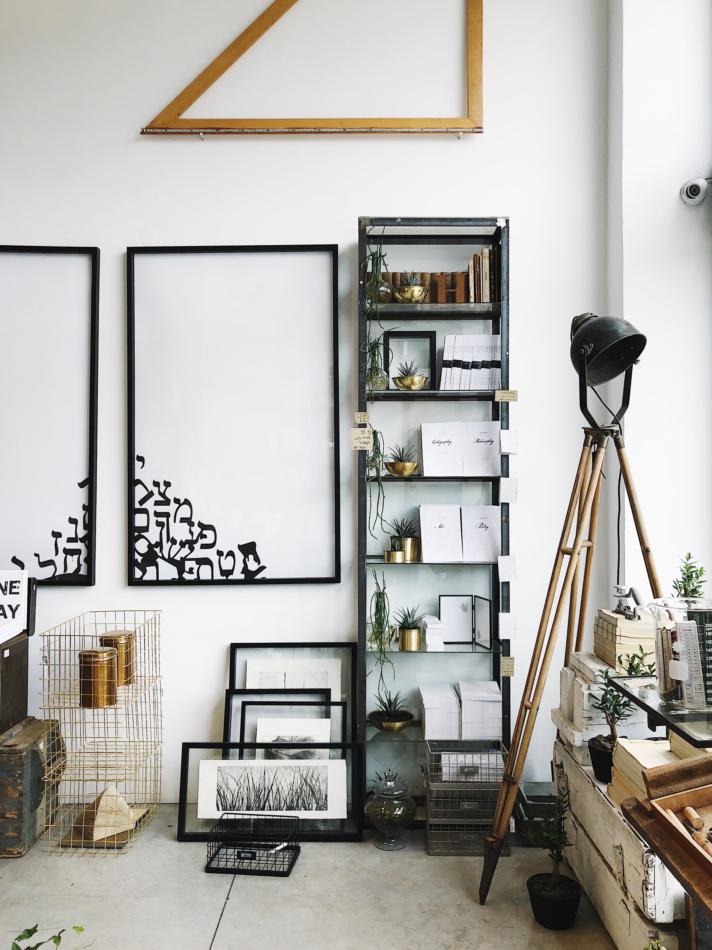 Interior of an art or home decor store with black picture frames, a metal shelving unit with books and small potted plants, a large vintage-style tripod lamp, and various decorative items including framed artwork, plants, jars, and a wire basket.