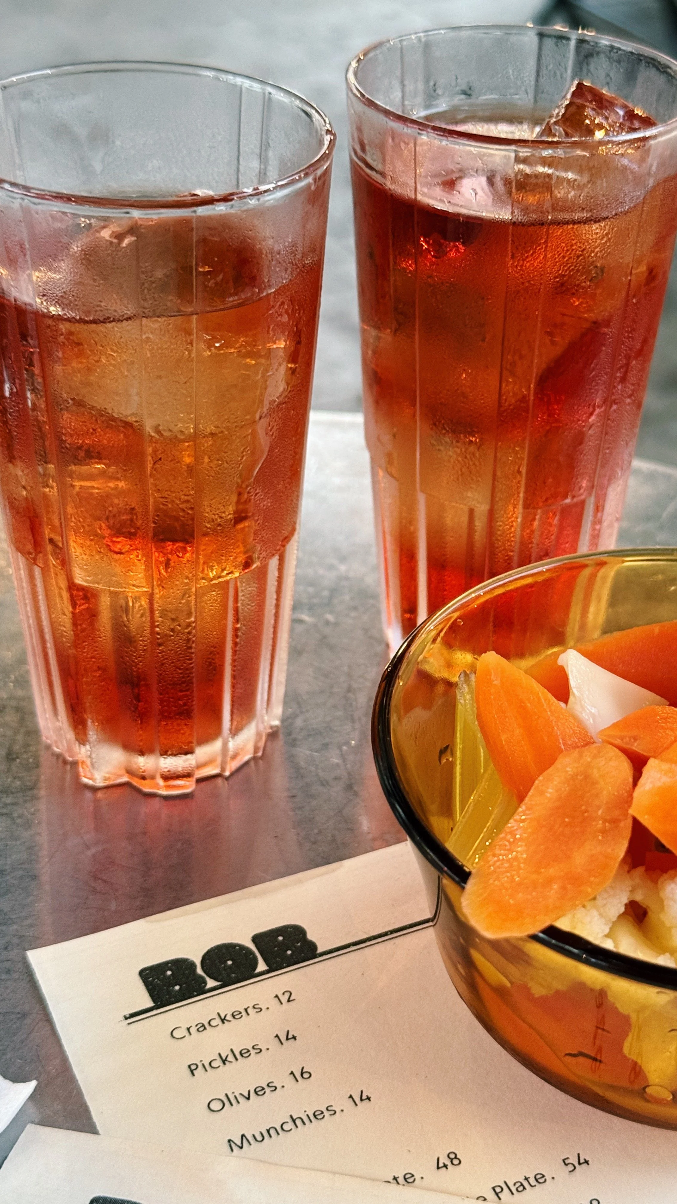 Two glasses of iced tea with ice cubes next to a bowl of pickled carrots and sundried vegetables on a table with a partially visible menu.