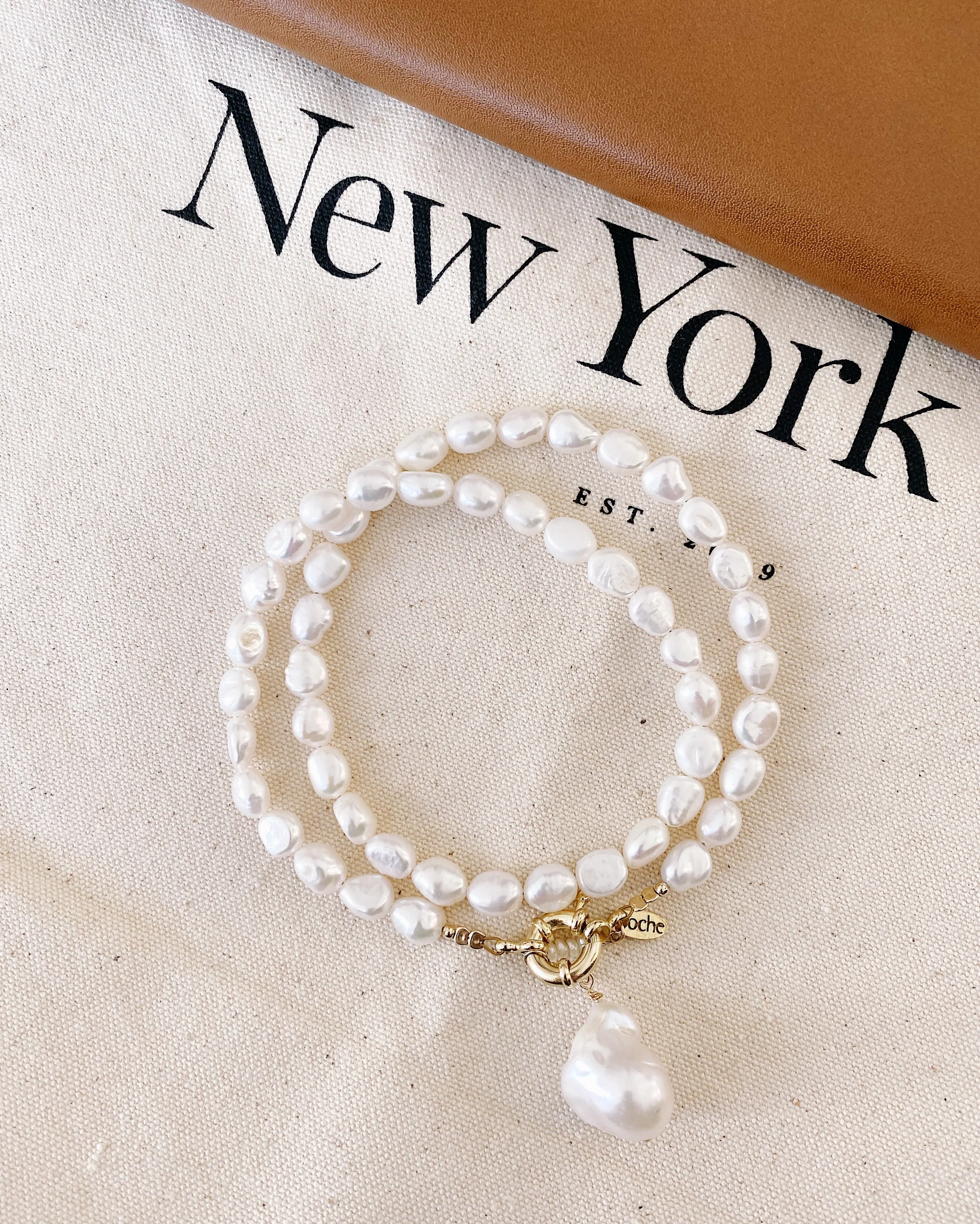 A pearl bracelet with a large pearl charm on a beige fabric surface next to a "New York" book.
