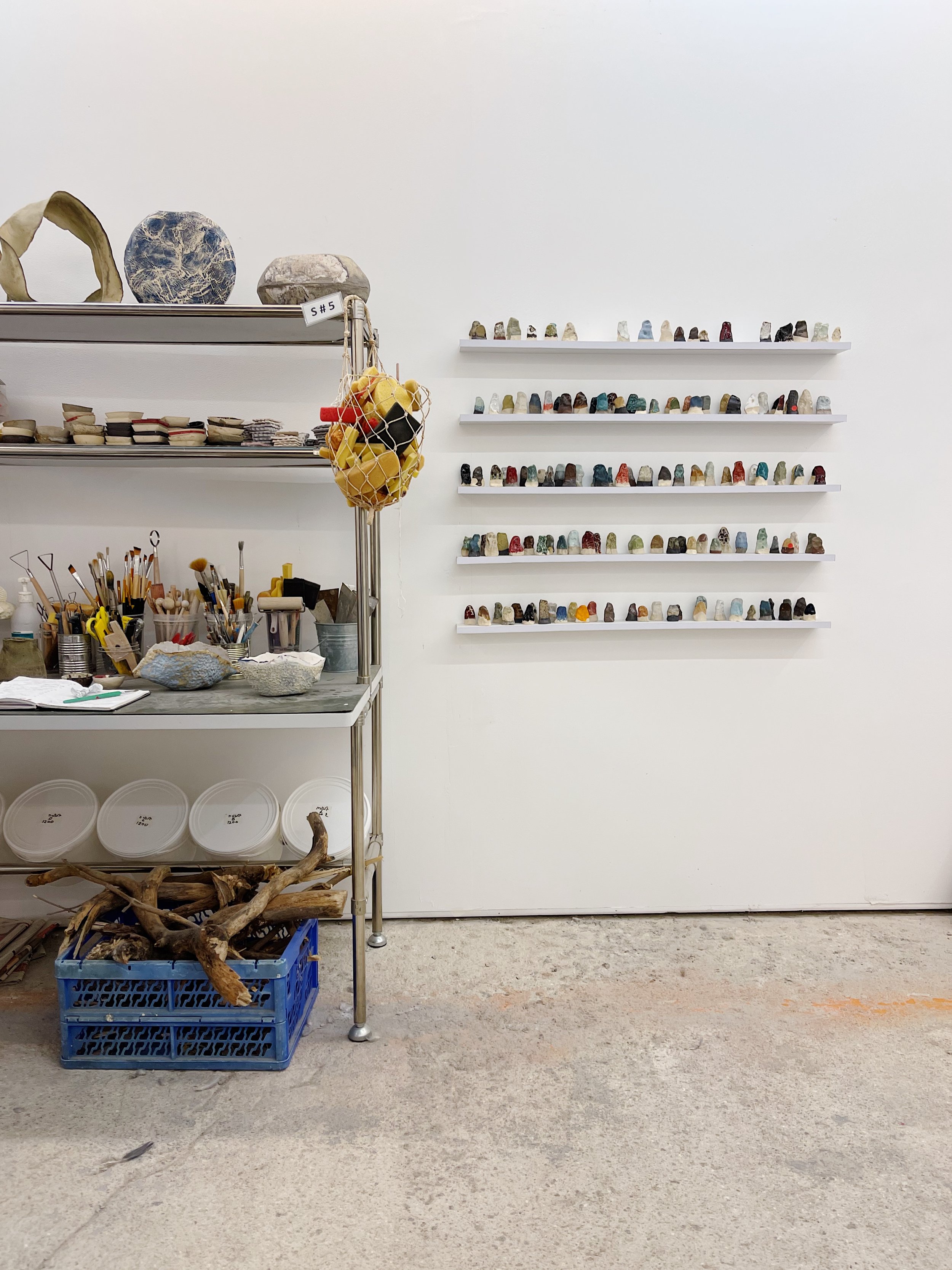 Display of small colorful stones on wall-mounted shelves in an artist's studio, with art supplies and rocks on a metal cart below.